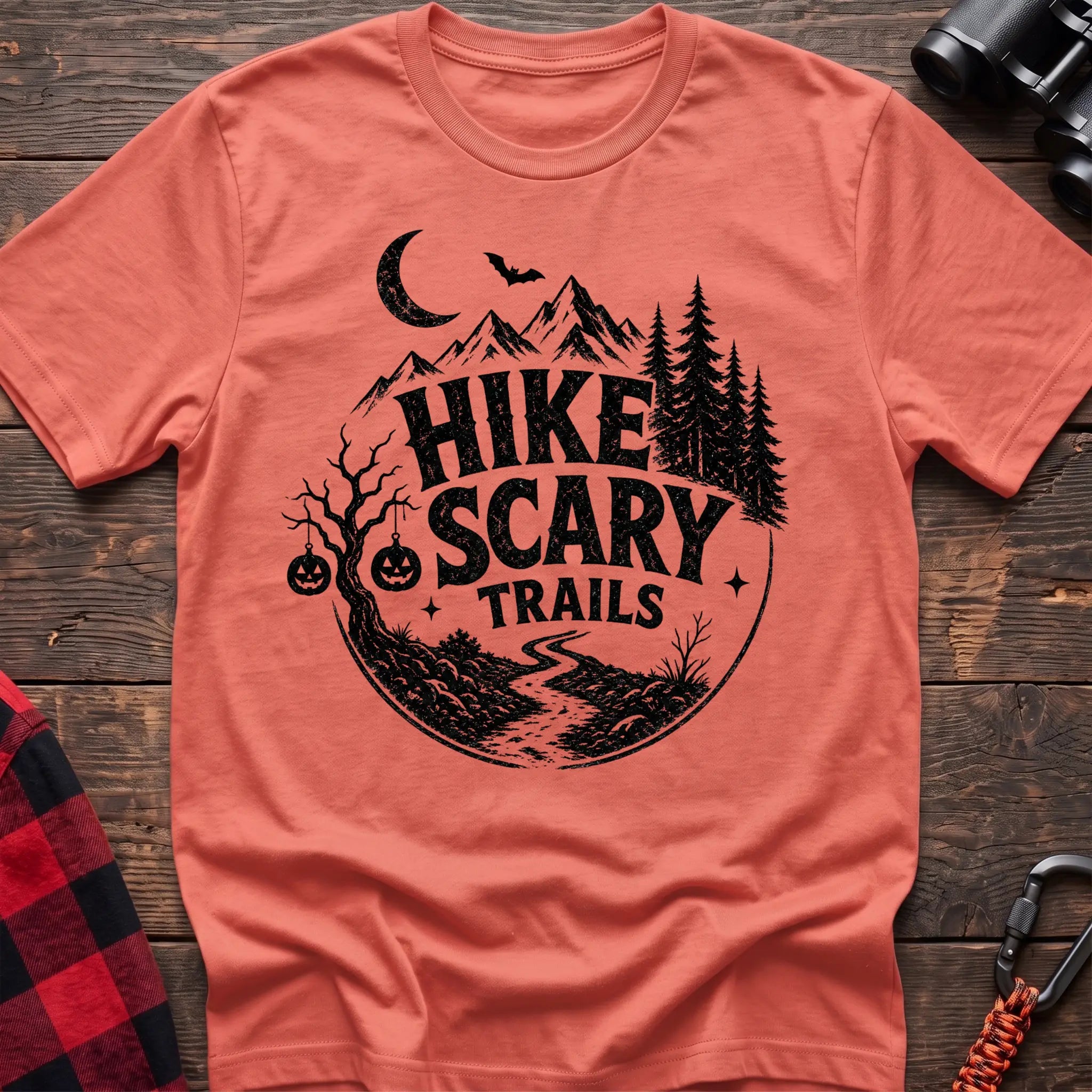 Heather orange cotton t-shirt with vintage Hike Scary Trails graphic of mountains, a crescent moon, bat, jack-o-lanterns, and a winding trail, flat lay on rustic wood with hiking gear