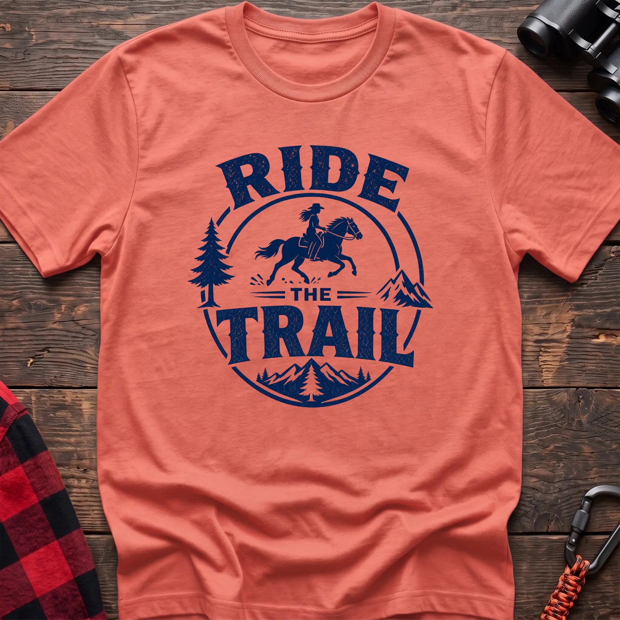 Heather orange cotton t-shirt with navy Ride the Trail graphic of a woman horseback riding through mountains and pine trees, flat lay on rustic wood with hiking gear