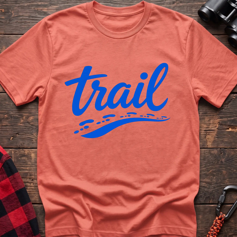 Heather orange cotton t-shirt with blue cursive trail script and dotted path graphic, flat lay on rustic wood with hiking gear