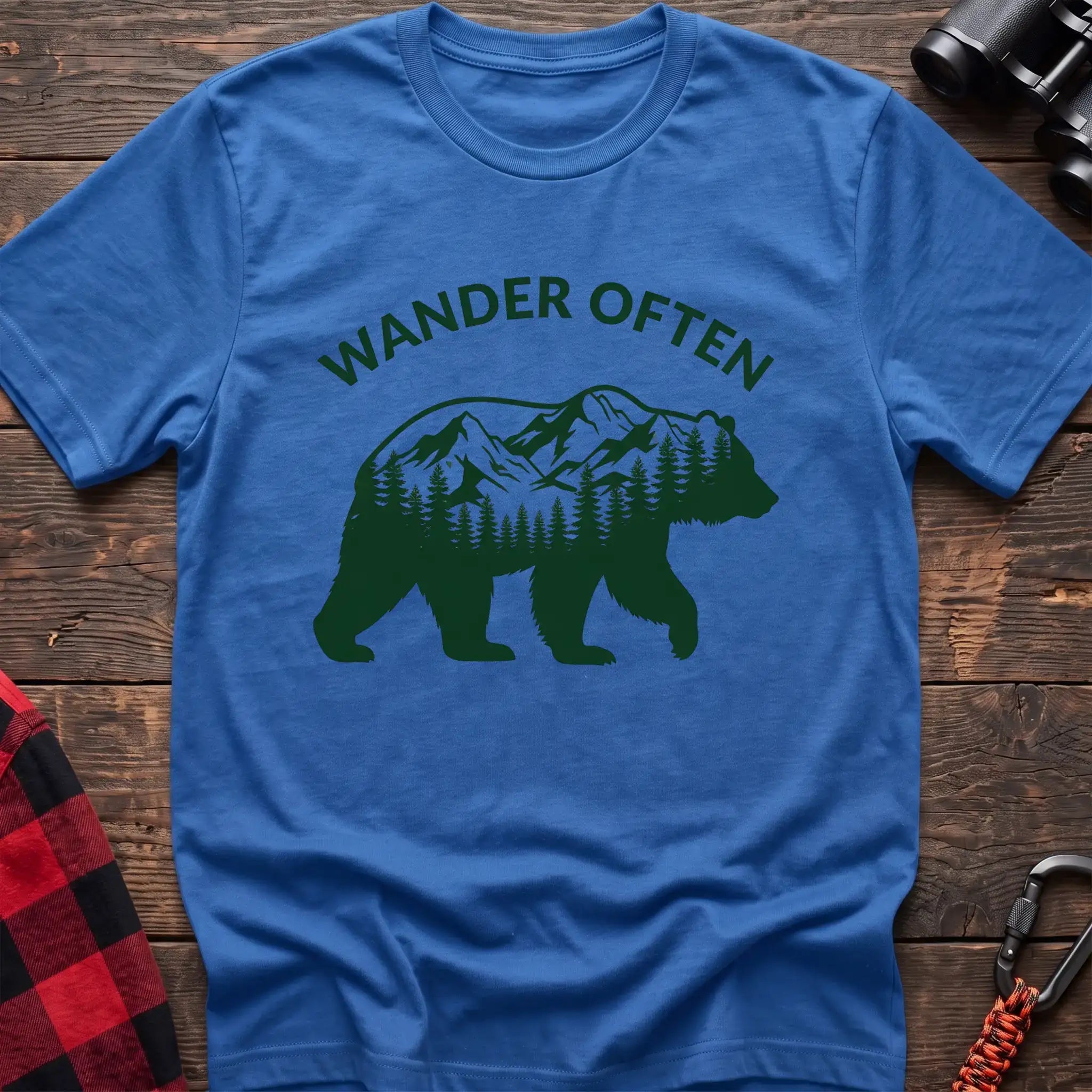 Heather royal t-shirt with a dark green silhouette of a walking bear containing a mountain and pine forest scene inside, with the arched text “WANDER OFTEN” above it, laid flat on a wooden surface.