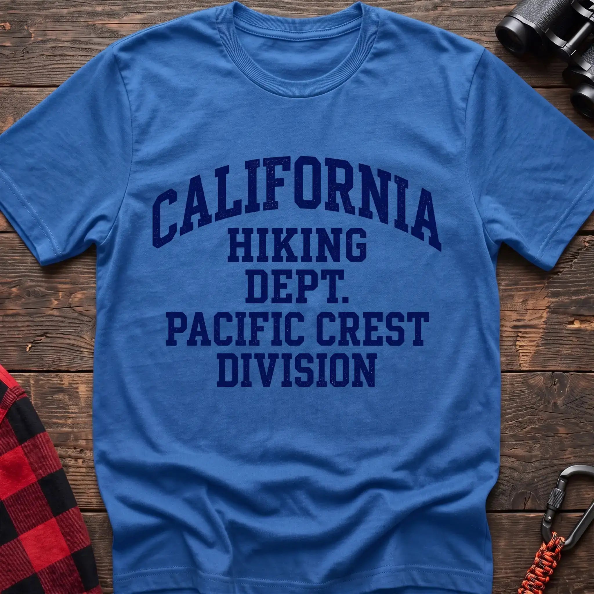 Heather royal t-shirt with distressed blue collegiate-style text reading “CALIFORNIA HIKING DEPT. PACIFIC CREST DIVISION,” laid flat on a wooden surface.