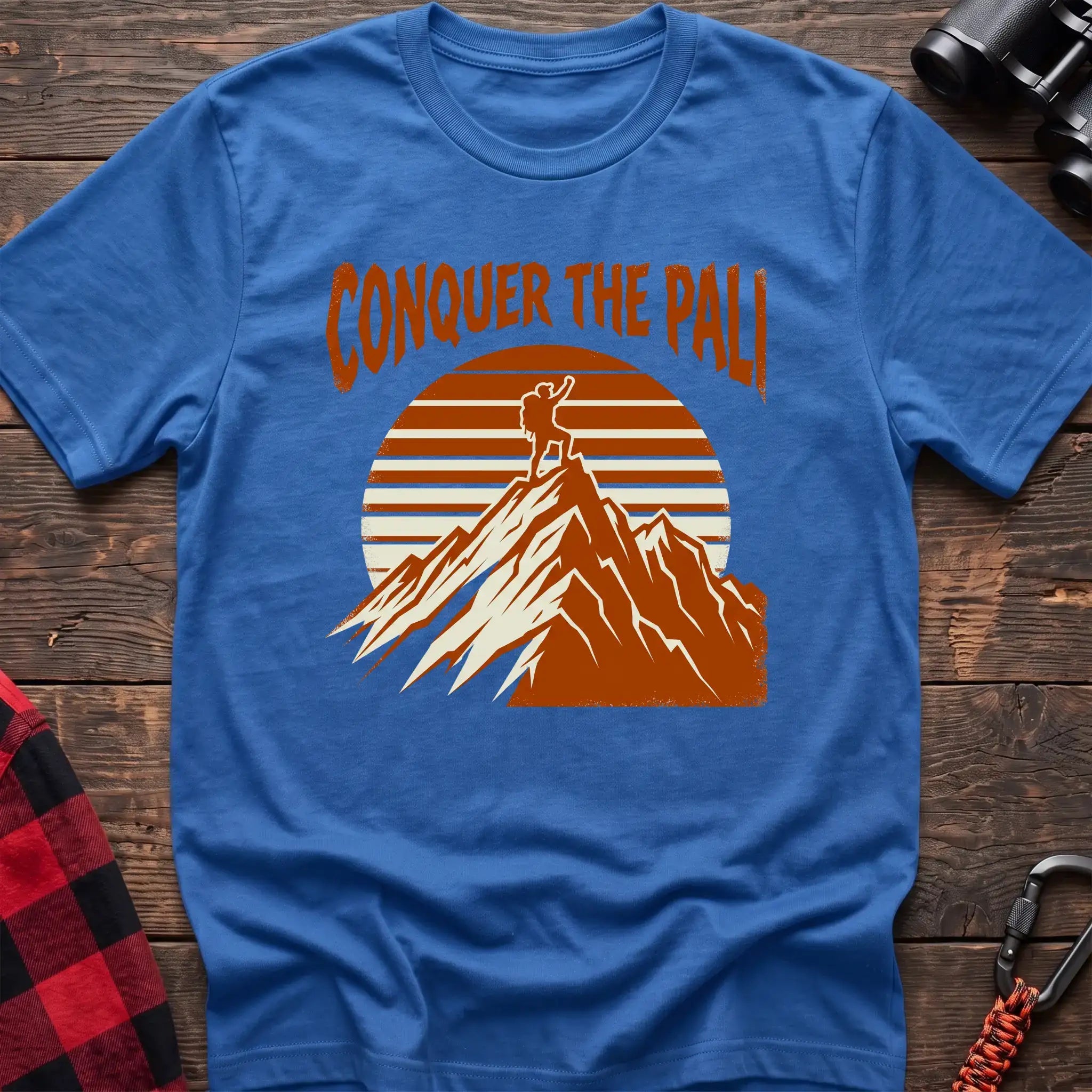 Heather royal t-shirt with orange text reading Conquer the Pali above a hiker standing on a mountain peak with a retro sun graphic