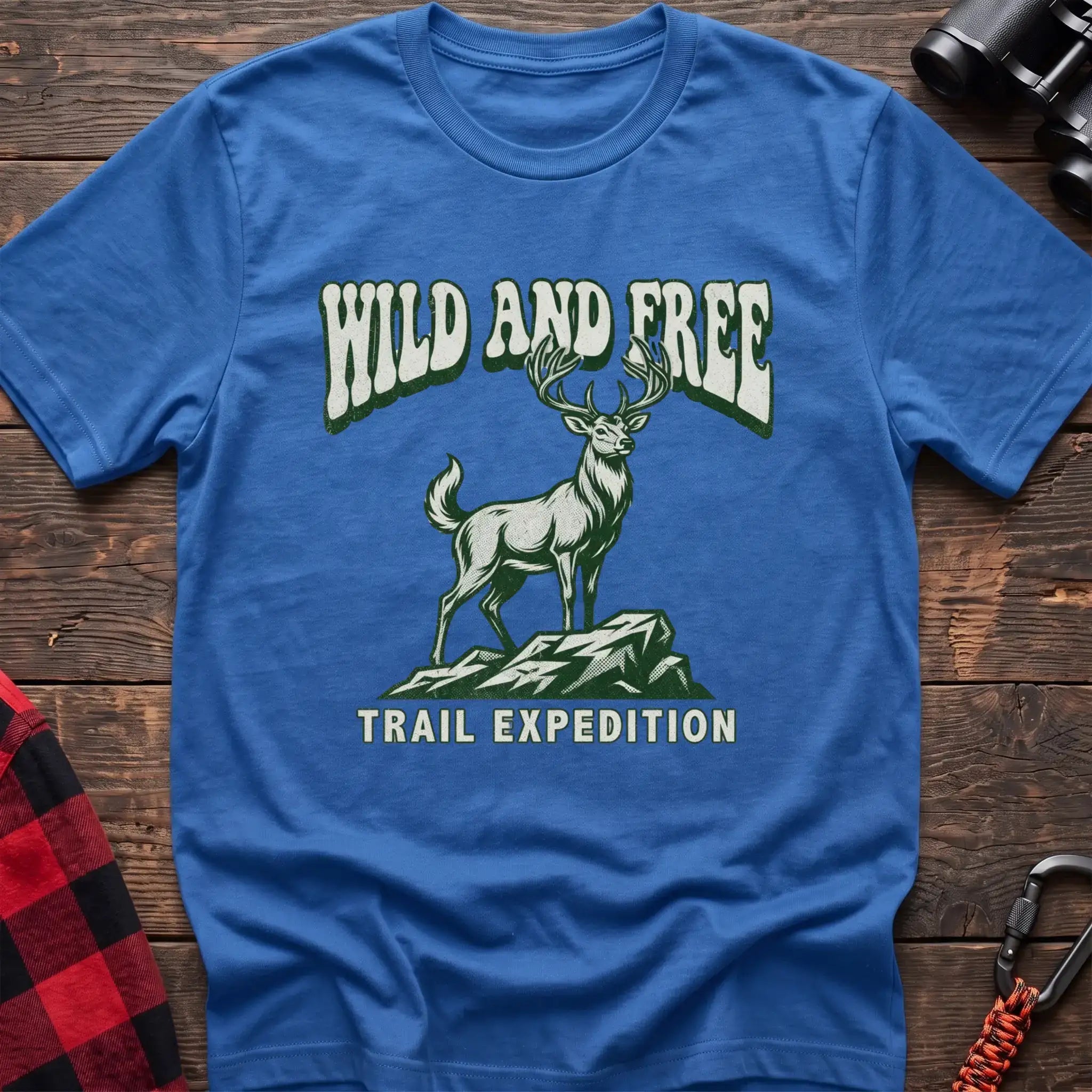 Heather royal t-shirt with a rustic graphic of a deer standing on a rocky outcrop, with the arched text “WILD AND FREE” above and “TRAIL EXPEDITION” below, laid flat on a wooden surface.
