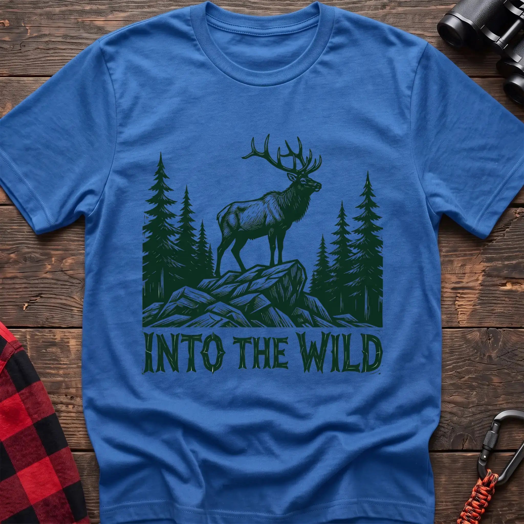 Heather royal t-shirt with a dark green illustration of an elk standing on a rocky outcrop among pine trees, with the text “INTO THE WILD” printed below, laid flat on a wooden surface.