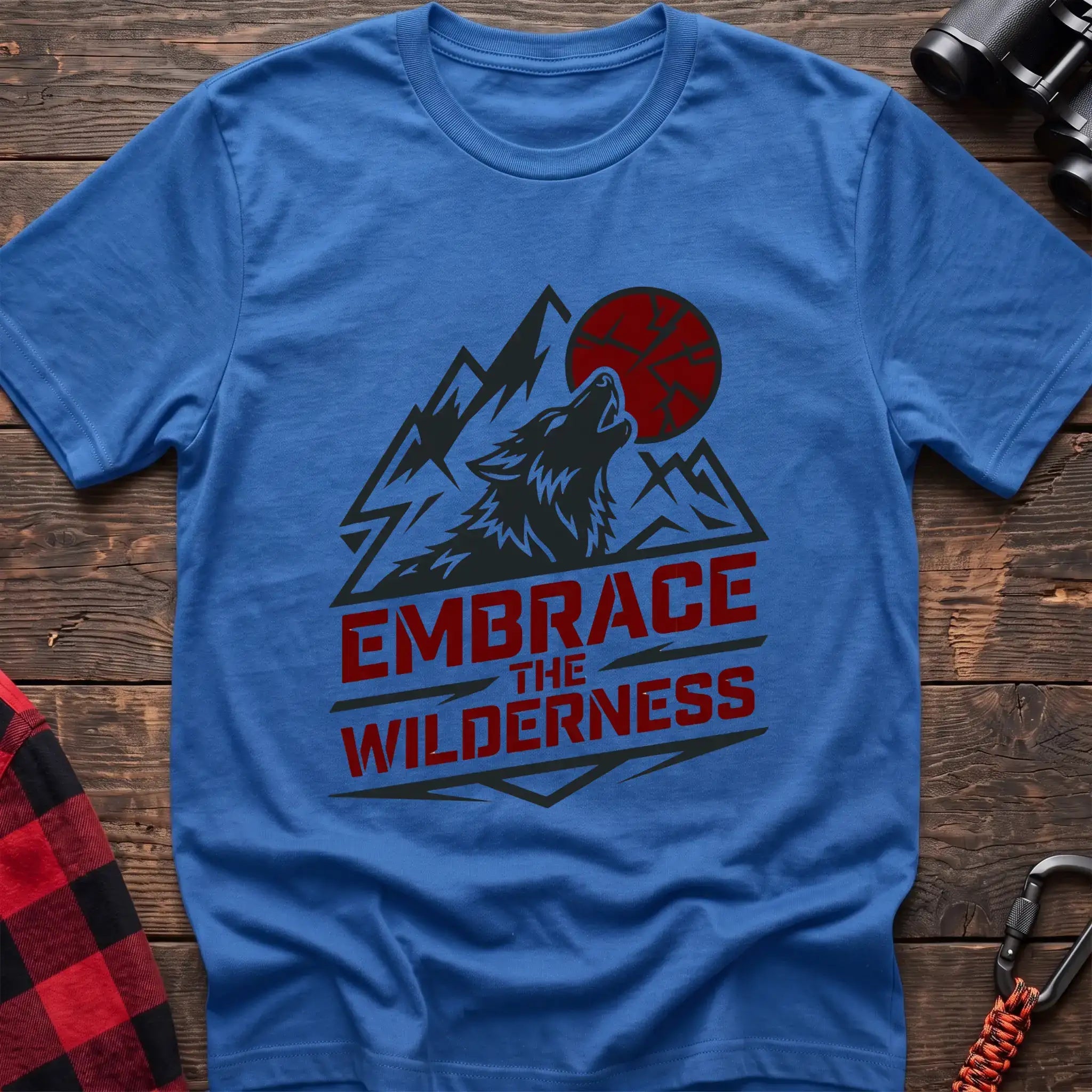 Heather royal t-shirt with a dark graphic of a howling wolf against mountain peaks and a red sun, with the bold red text “EMBRACE THE WILDERNESS” below, laid flat on a wooden surface.