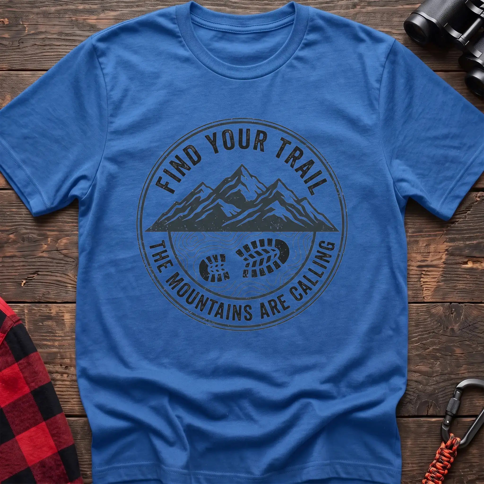 Heather royal t-shirt with a circular dark gray badge design featuring mountain peaks above a hiking boot print on topographic lines, with the text “FIND YOUR TRAIL” and “THE MOUNTAINS ARE CALLING,” laid flat on a wooden surface.