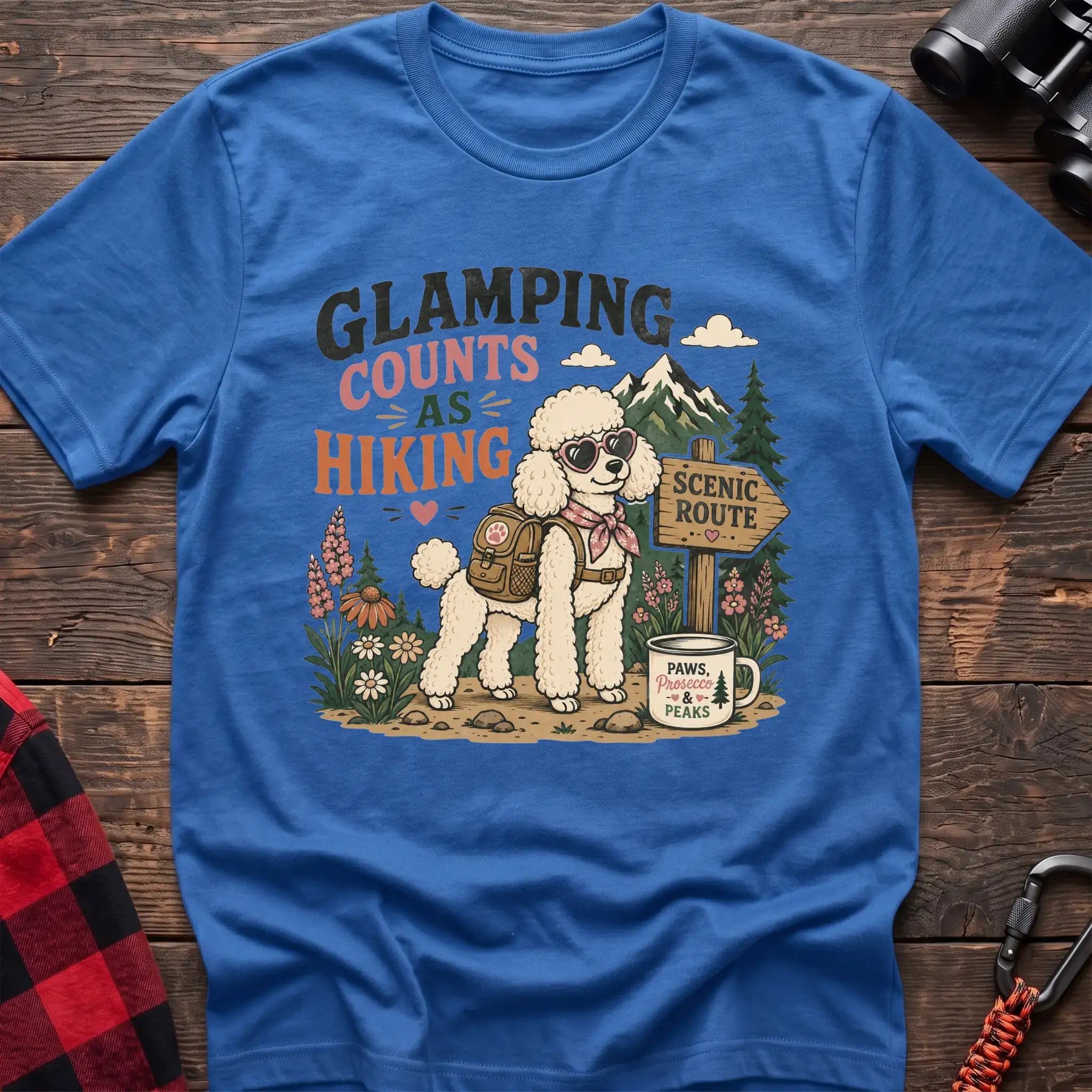 Heather royal t-shirt with a colorful illustration of a poodle in sunglasses and a backpack on a scenic trail, with the text “GLAMPING COUNTS AS HIKING,” laid flat on a wooden surface.