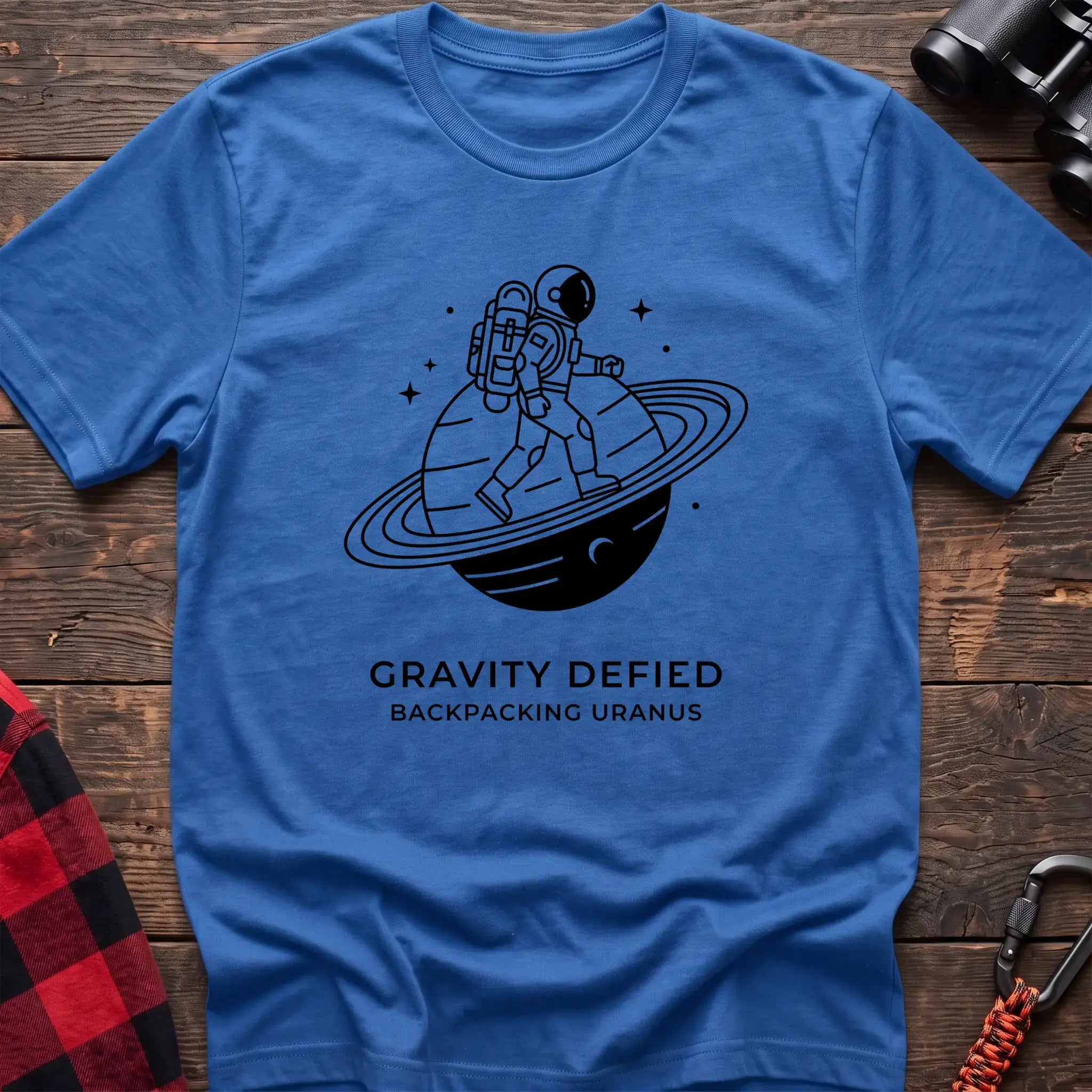 Heather royal crew-neck t-shirt with black line-art graphic of an astronaut backpacking across a ringed planet surrounded by stars, with the text “GRAVITY DEFIED / BACKPACKING URANUS” printed below, laid flat on a wooden surface.