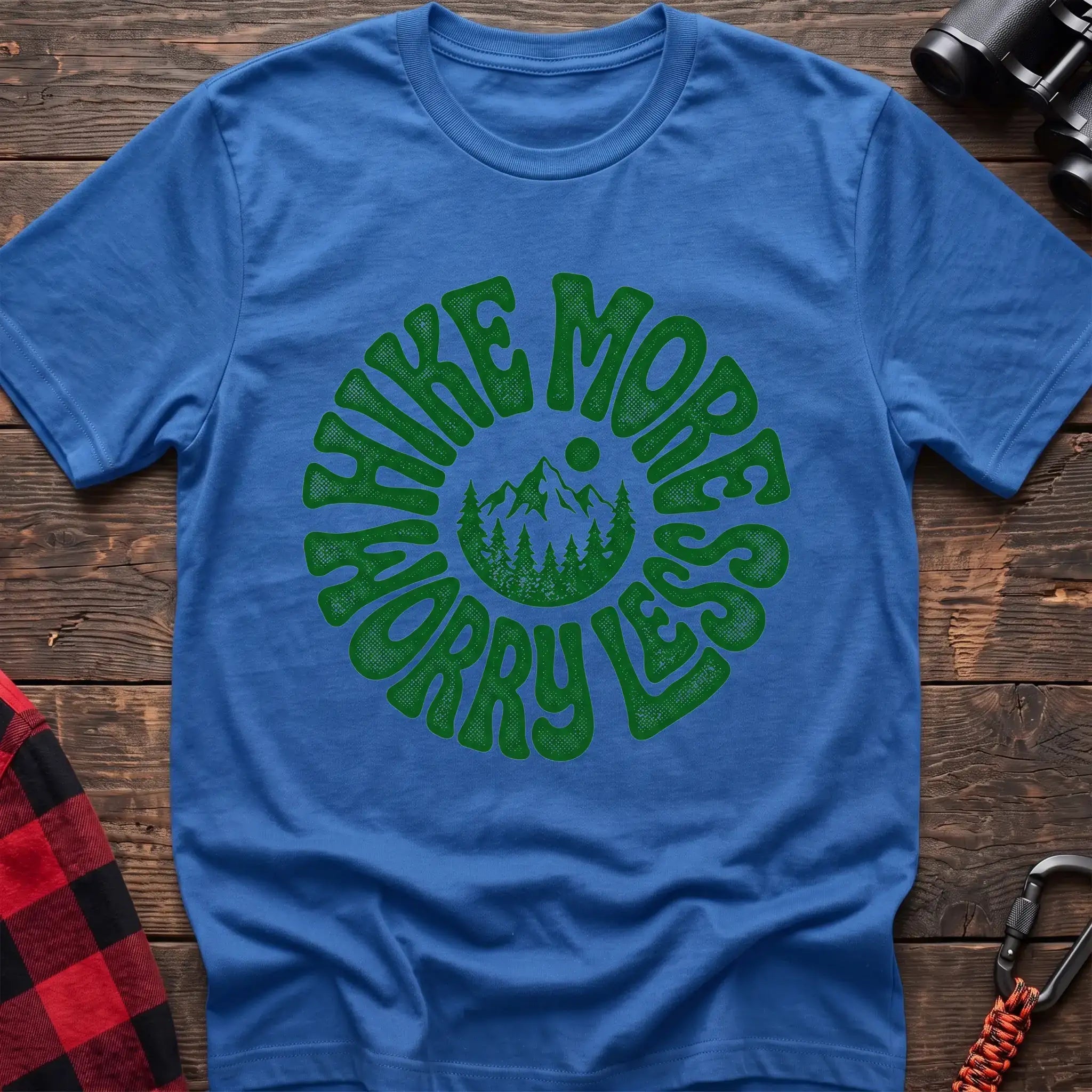 Heather royal t-shirt with a circular green design featuring mountains and pine trees in the center, surrounded by the text “HIKE MORE WORRY LESS,” laid flat on a wooden surface.