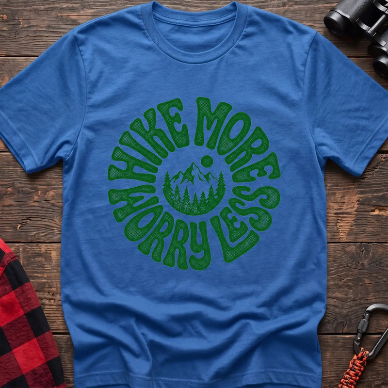 Heather royal t-shirt with a circular green design featuring mountains and pine trees in the center, surrounded by the text “HIKE MORE WORRY LESS,” laid flat on a wooden surface.
