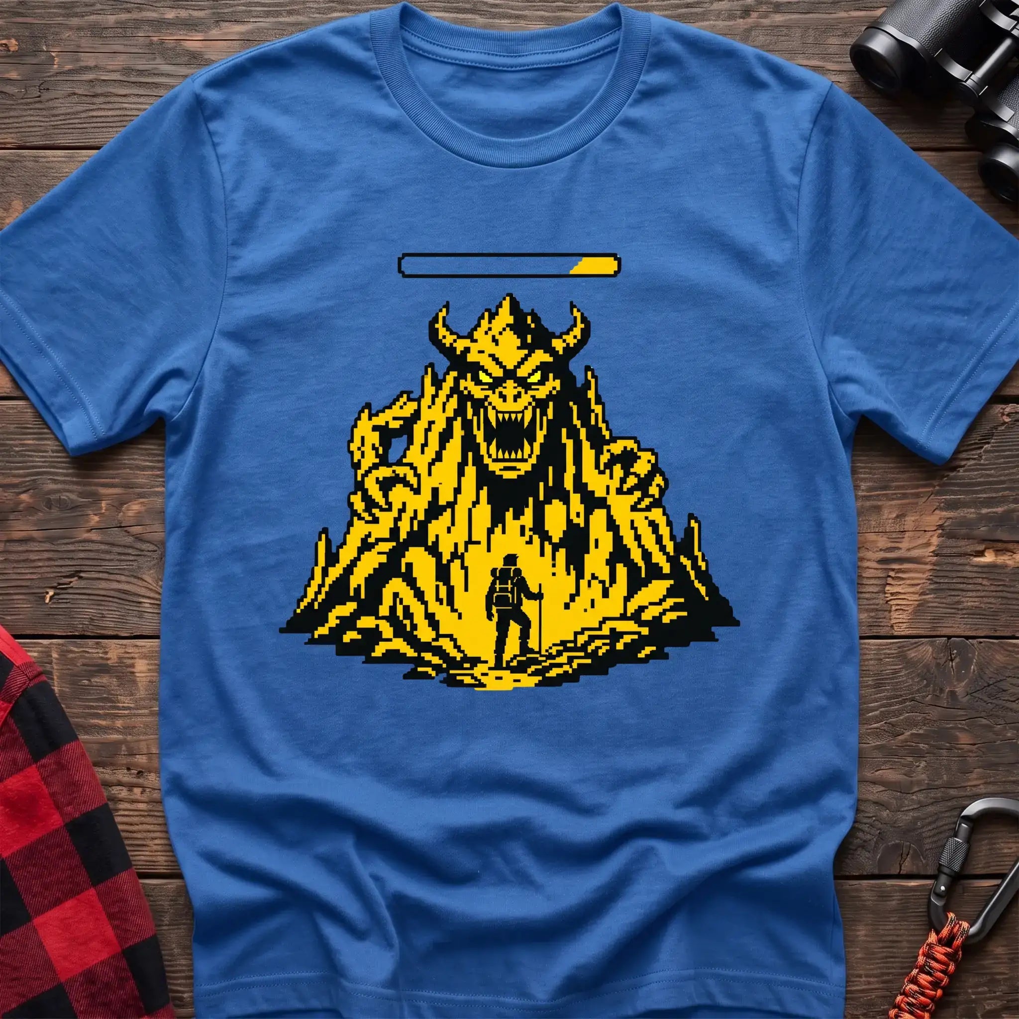 Heather royal t-shirt with a pixel-art illustration of a hiker facing a large horned mountain monster with a health bar above it, laid flat on a wooden surface.