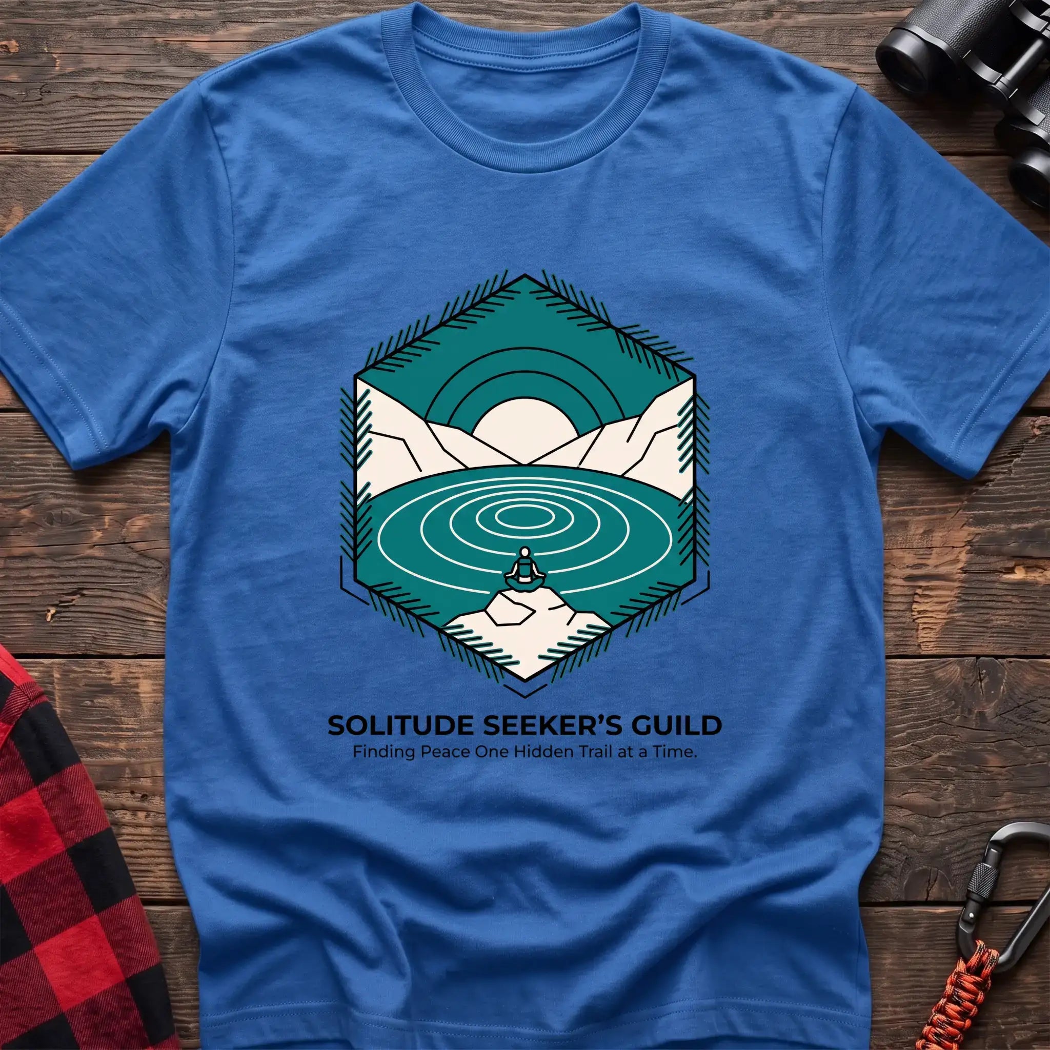 Heather royal t-shirt with a hexagonal illustration of a person meditating by a mountain lake, with the text “SOLITUDE SEEKER’S GUILD” and “Finding Peace One Hidden Trail at a Time,” laid flat on a wooden surface.