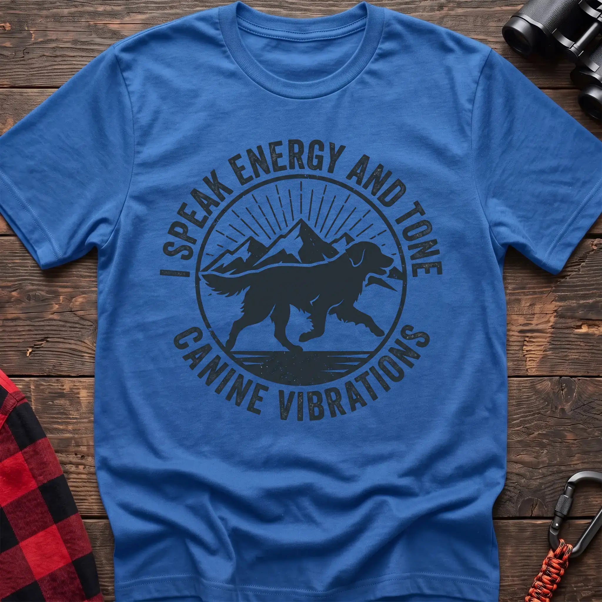 Heather royal t-shirt with a circular dark gray badge design featuring a running dog carrying a stick against mountain peaks and sun rays, with the text “I SPEAK ENERGY AND TONE” and “CANINE VIBRATIONS,” laid flat on a wooden surface.