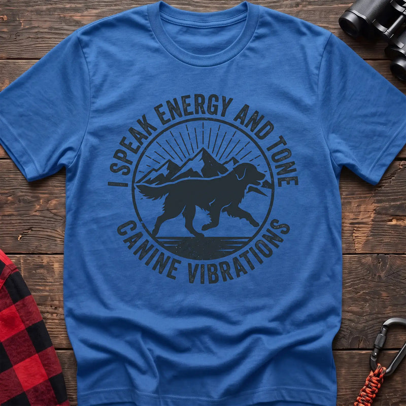 Heather royal t-shirt with a circular dark gray badge design featuring a running dog carrying a stick against mountain peaks and sun rays, with the text “I SPEAK ENERGY AND TONE” and “CANINE VIBRATIONS,” laid flat on a wooden surface.