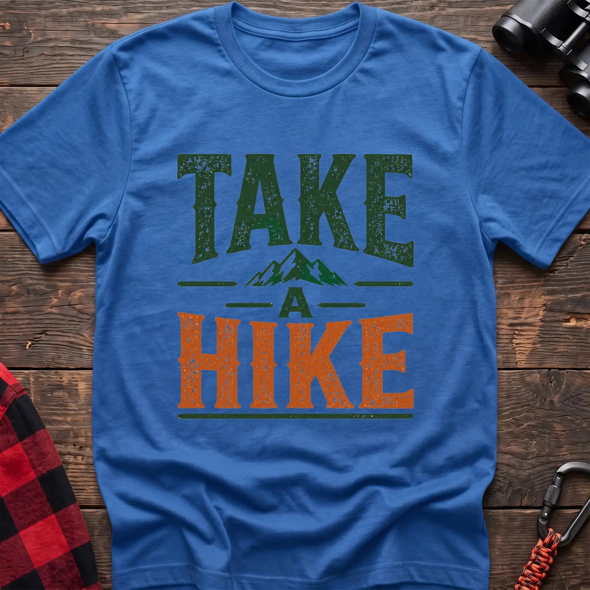 Heather royal t-shirt with distressed green and orange text reading “TAKE A HIKE” with a small mountain graphic between the words, laid flat on a wooden surface.