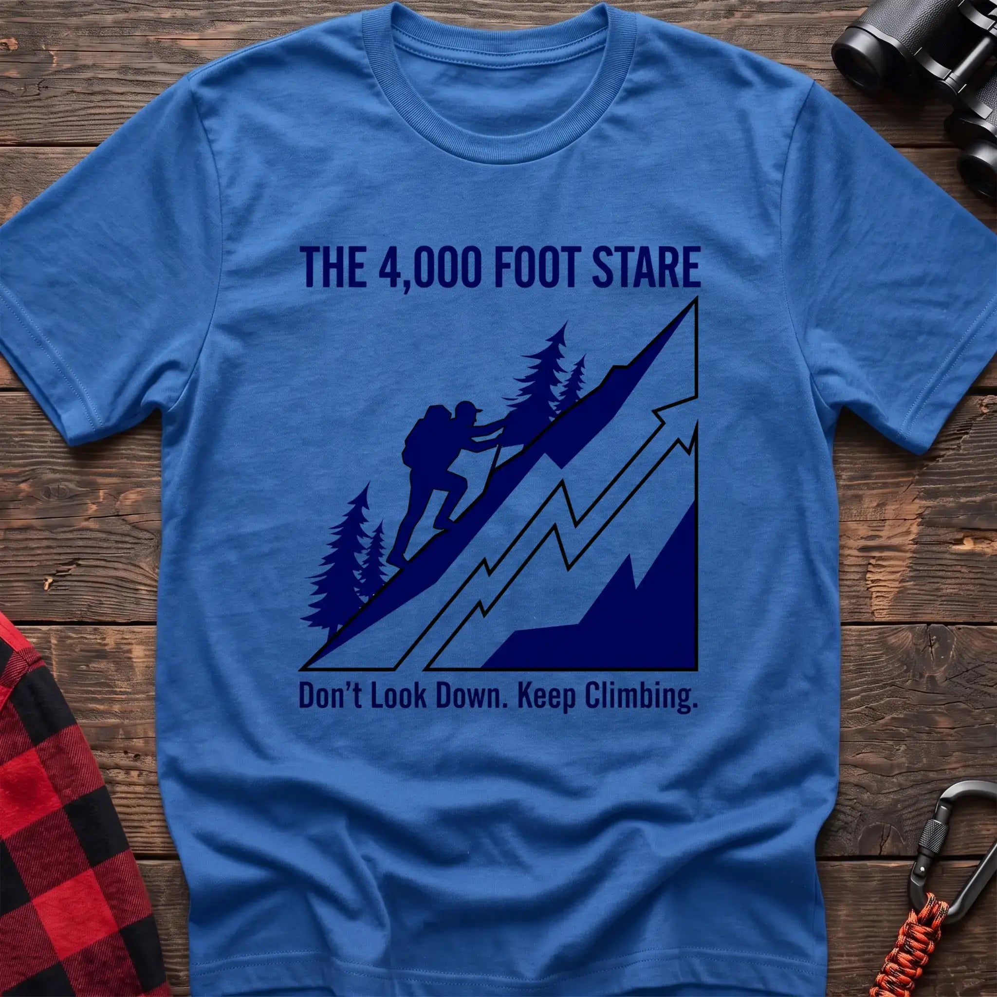 Heather royal t-shirt with a blue illustration of a hiker climbing a steep mountain, with the text “THE 4,000 FOOT STARE” and “Don’t Look Down. Keep Climbing,” laid flat on a wooden surface.