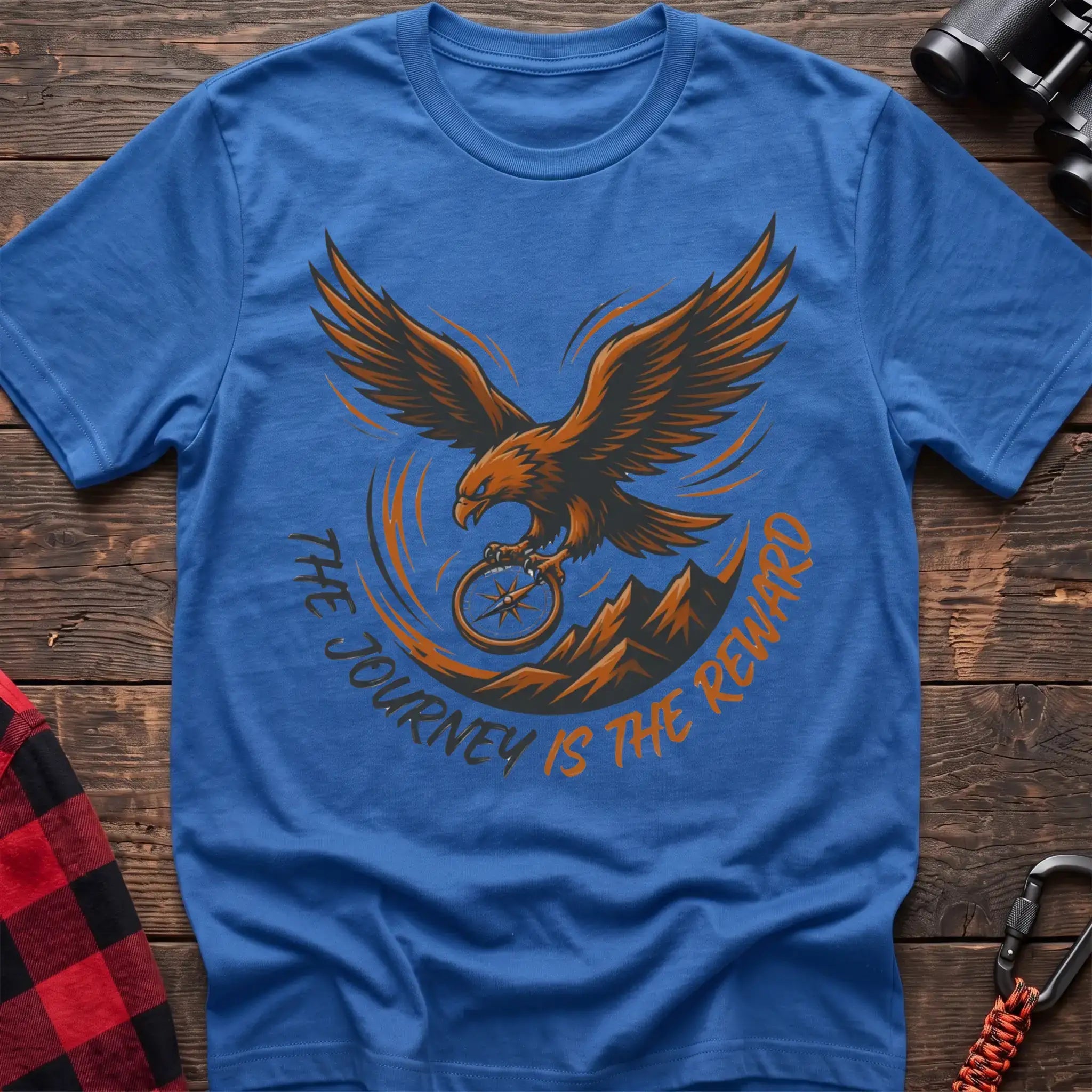 Heather royal t-shirt with an orange and dark graphic of a soaring eagle holding a compass above mountains, with the curved text “THE JOURNEY IS THE REWARD,” laid flat on a wooden surface.