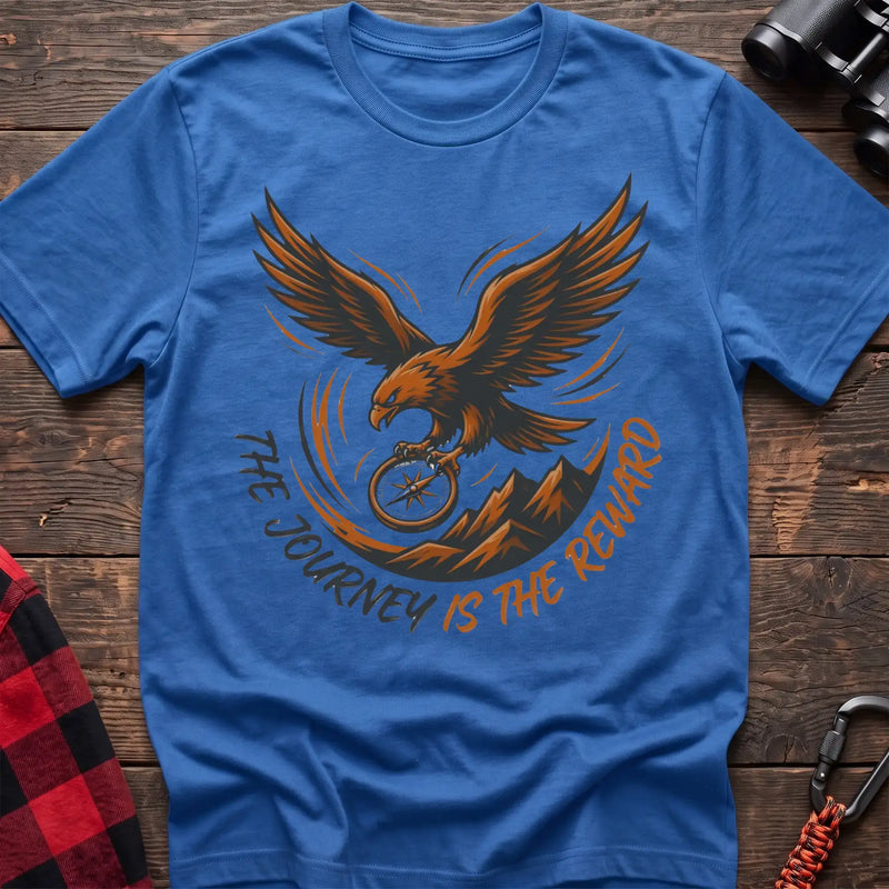 Heather royal t-shirt with an orange and dark graphic of a soaring eagle holding a compass above mountains, with the curved text “THE JOURNEY IS THE REWARD,” laid flat on a wooden surface.