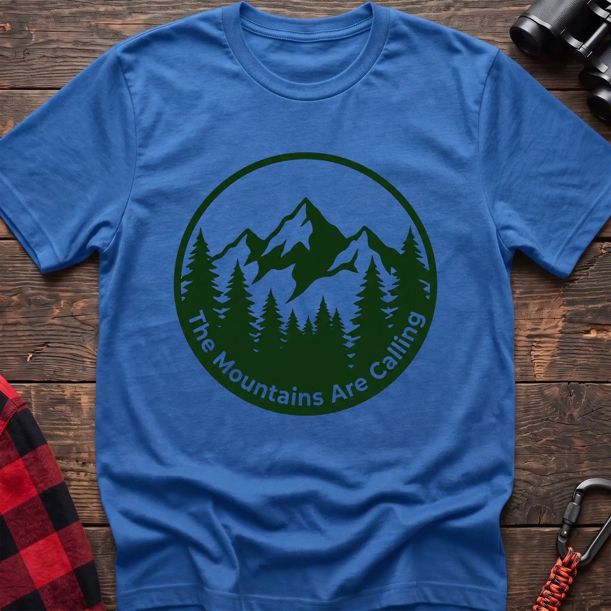 Heather royal t-shirt with a circular dark green design featuring mountain peaks and pine trees, with the text “The Mountains Are Calling,” laid flat on a wooden surface.