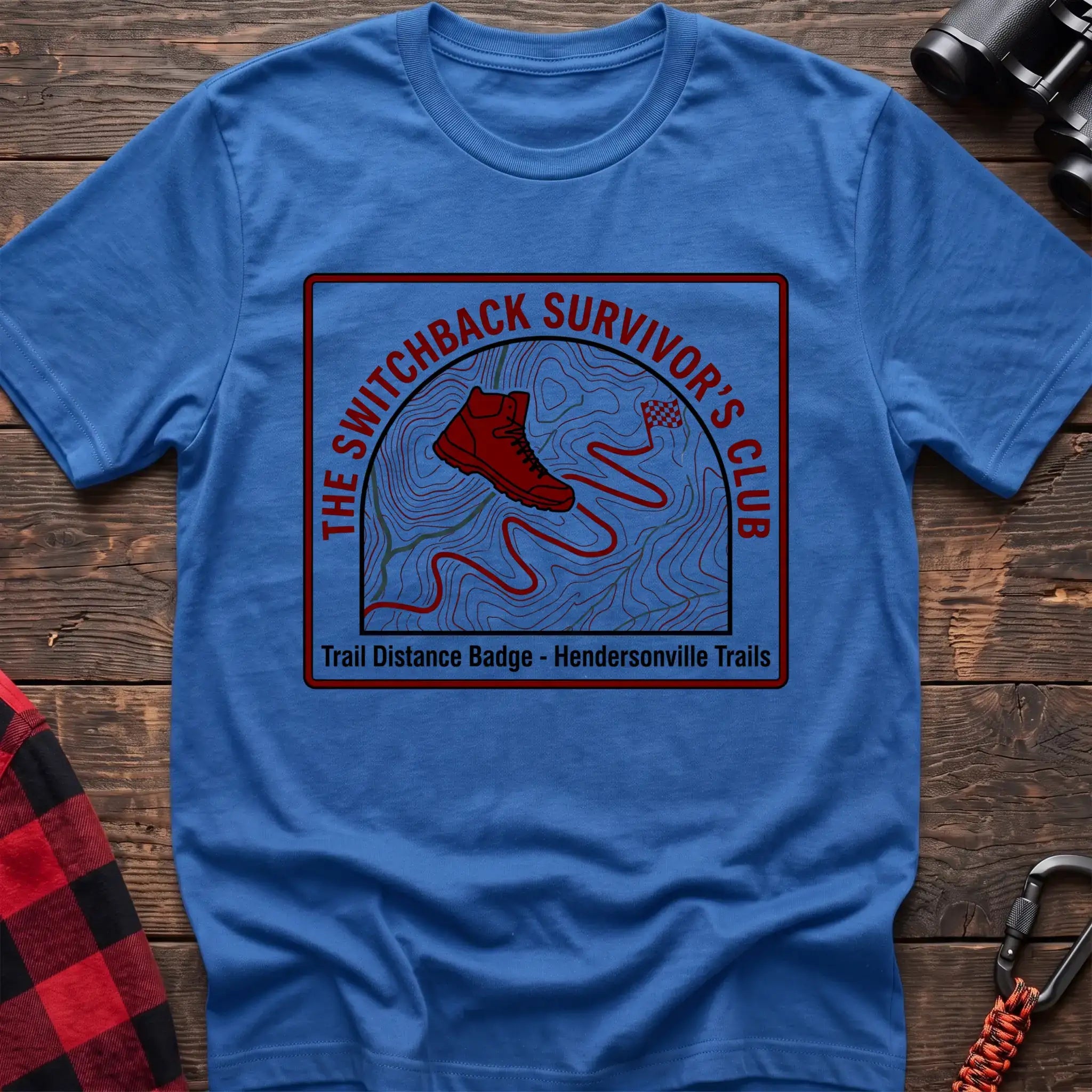 Heather royal t-shirt with a square badge design featuring a red hiking boot on a topographic map with a winding trail, and the text “THE SWITCHBACK SURVIVOR’S CLUB” and “Trail Distance Badge - Hendersonville Trails,” laid flat on a wooden surface.