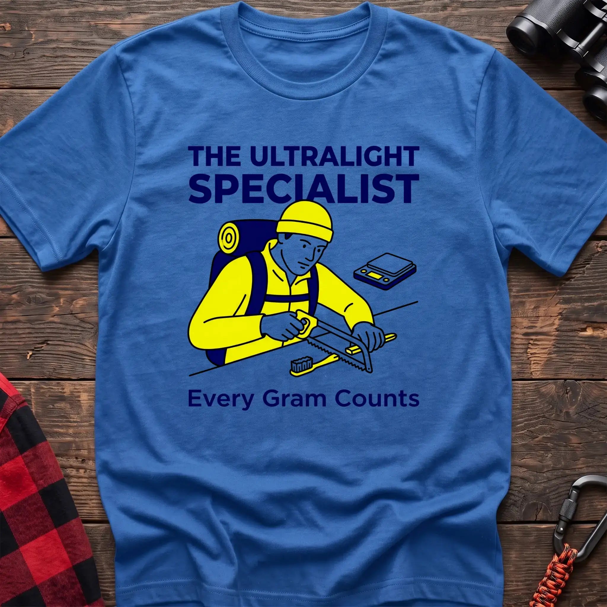 Heather royal t-shirt with an illustration of a hiker sawing a toothbrush on a table next to a scale, with the text “THE ULTRALIGHT SPECIALIST” and “Every Gram Counts,” laid flat on a wooden surface.