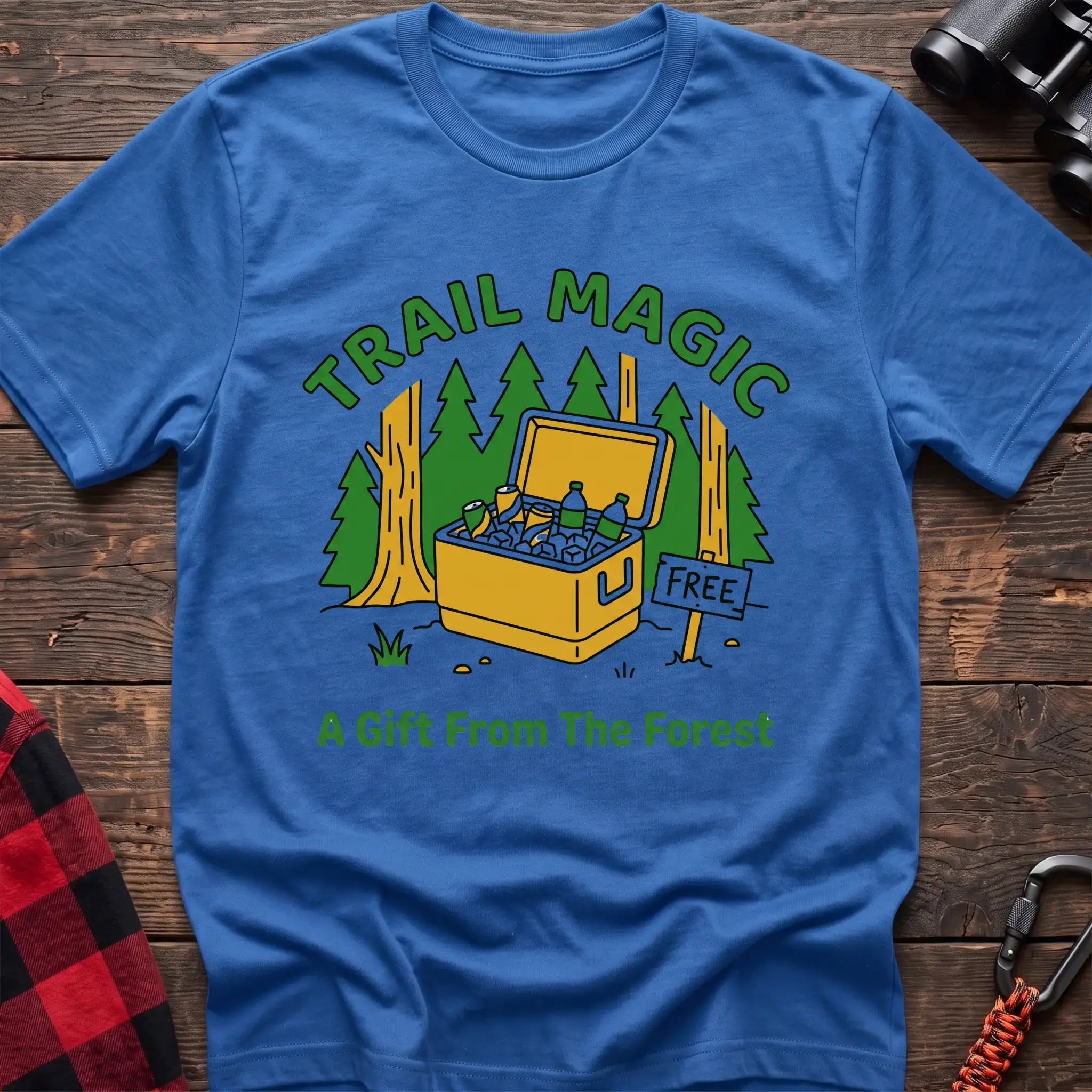 Heather royal t-shirt with an illustration of an open cooler filled with drinks in a forest setting, with the text “TRAIL MAGIC” and “A Gift From The Forest,” laid flat on a wooden surface.