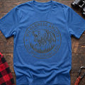 Heather royal t-shirt with a circular dark gray badge design featuring a roaring bear head above mountains and pine trees, with the text “WILDERNESS AWAITS” and “EST. 1974 - EXPLORE MORE,” laid flat on a wooden surface.