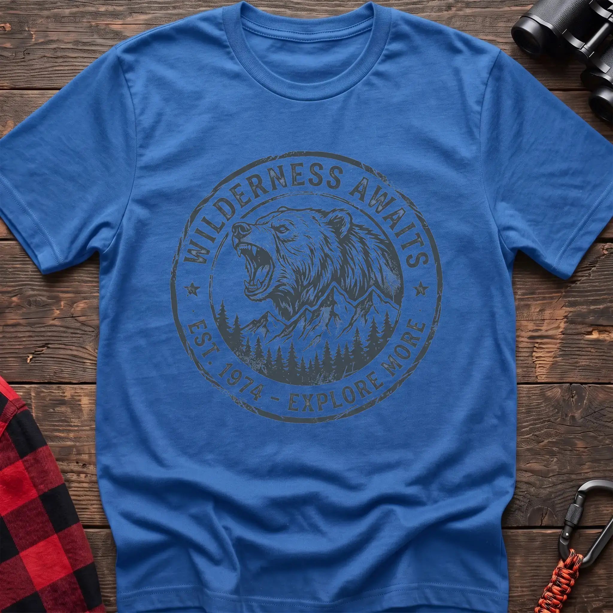 Heather royal t-shirt with a circular dark gray badge design featuring a roaring bear head above mountains and pine trees, with the text “WILDERNESS AWAITS” and “EST. 1974 - EXPLORE MORE,” laid flat on a wooden surface.