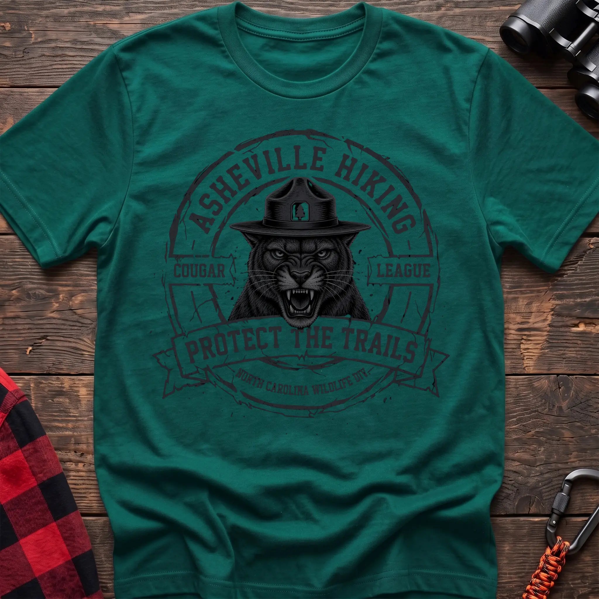 Kelly green cotton t-shirt with vintage Asheville Hiking Cougar League graphic of a roaring cougar in a park ranger hat, Protect the Trails North Carolina Wildlife Division badge, flat lay on rustic wood with hiking gear