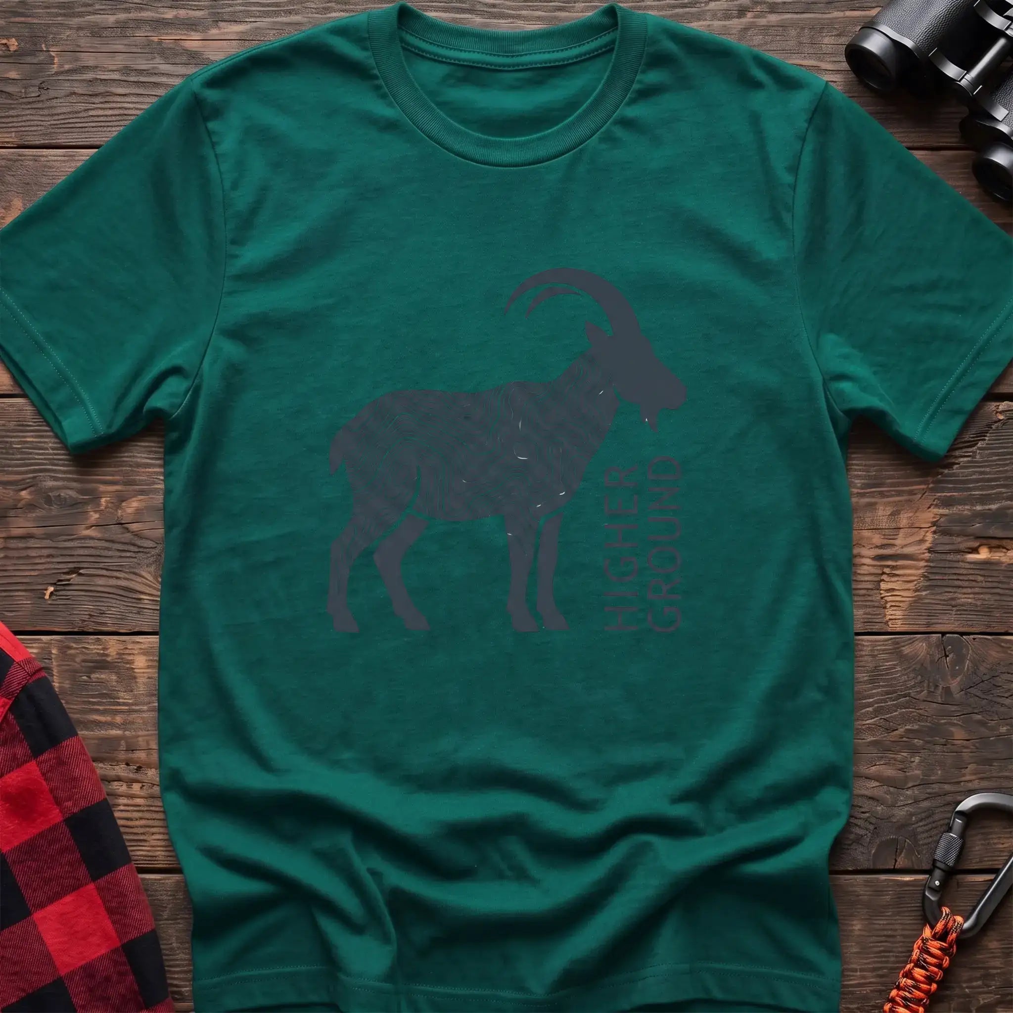 Kelly green t-shirt with a dark silhouette of a mountain goat filled with topographic contour lines, and the vertical text “HIGHER GROUND” printed beside it, laid flat on a wooden surface.