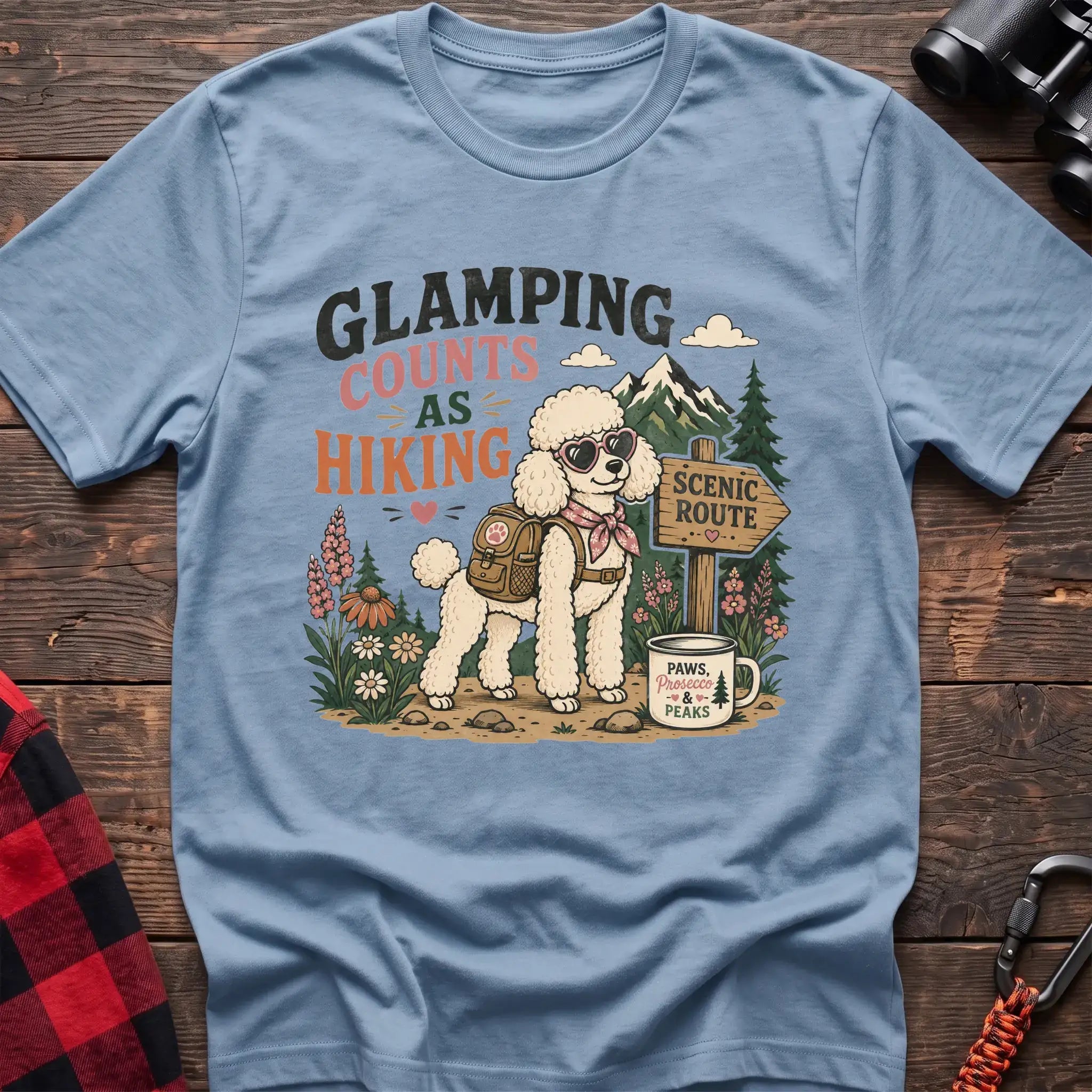 Light Blue t-shirt with a colorful illustration of a poodle in sunglasses and a backpack on a scenic trail, with the text “GLAMPING COUNTS AS HIKING,” laid flat on a wooden surface.