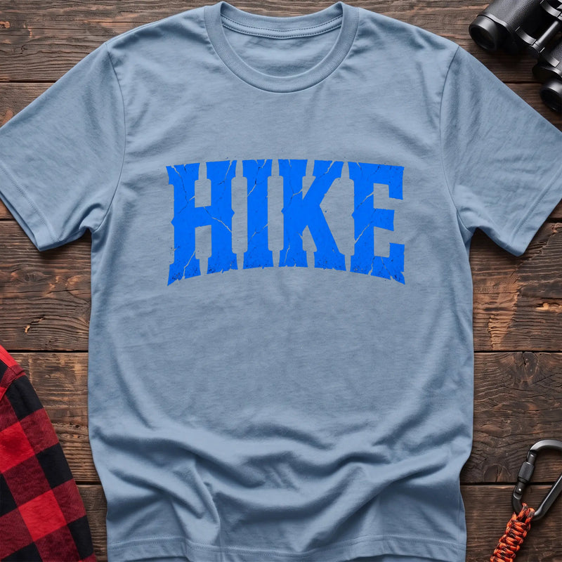 Light blue cotton t-shirt with distressed arched blue HIKE graphic in cracked varsity lettering, flat lay on rustic wood with hiking gear