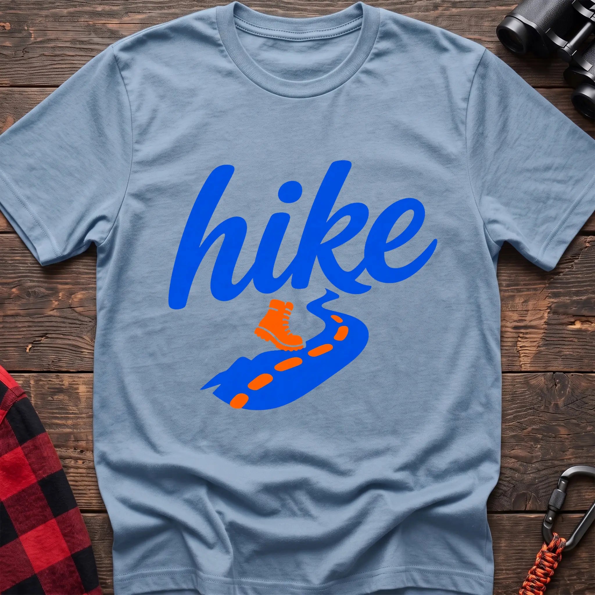 Light blue cotton t-shirt with royal blue cursive hike script, orange hiking boot, and blue trail with orange footprints, flat lay on rustic wood with hiking gear