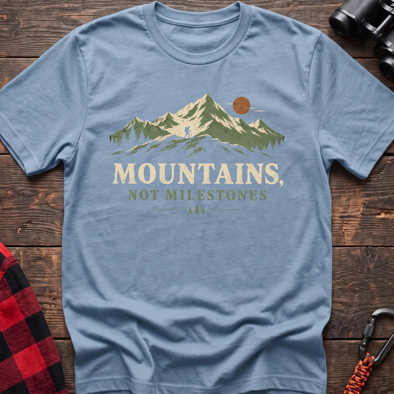 Light blue cotton t-shirt with a hiker and mountain graphic and Mountains Not Milestones text on rustic wood