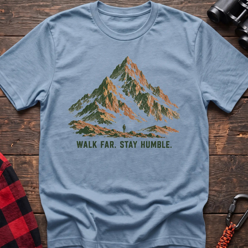 Light blue cotton t-shirt with a mountain and hiker graphic and Walk Far Stay Humble text on rustic wood