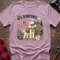 Light Pink t-shirt with a colorful illustration of a poodle in sunglasses and a backpack on a scenic trail, with the text “GLAMPING COUNTS AS HIKING,” laid flat on a wooden surface.