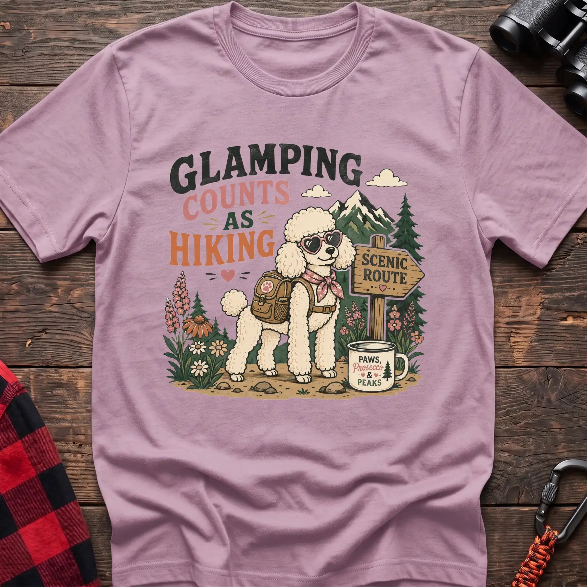 Light Pink t-shirt with a colorful illustration of a poodle in sunglasses and a backpack on a scenic trail, with the text “GLAMPING COUNTS AS HIKING,” laid flat on a wooden surface.