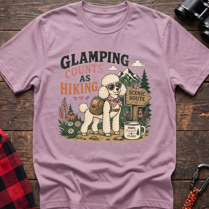 Light Pink t-shirt with a colorful illustration of a poodle in sunglasses and a backpack on a scenic trail, with the text “GLAMPING COUNTS AS HIKING,” laid flat on a wooden surface.