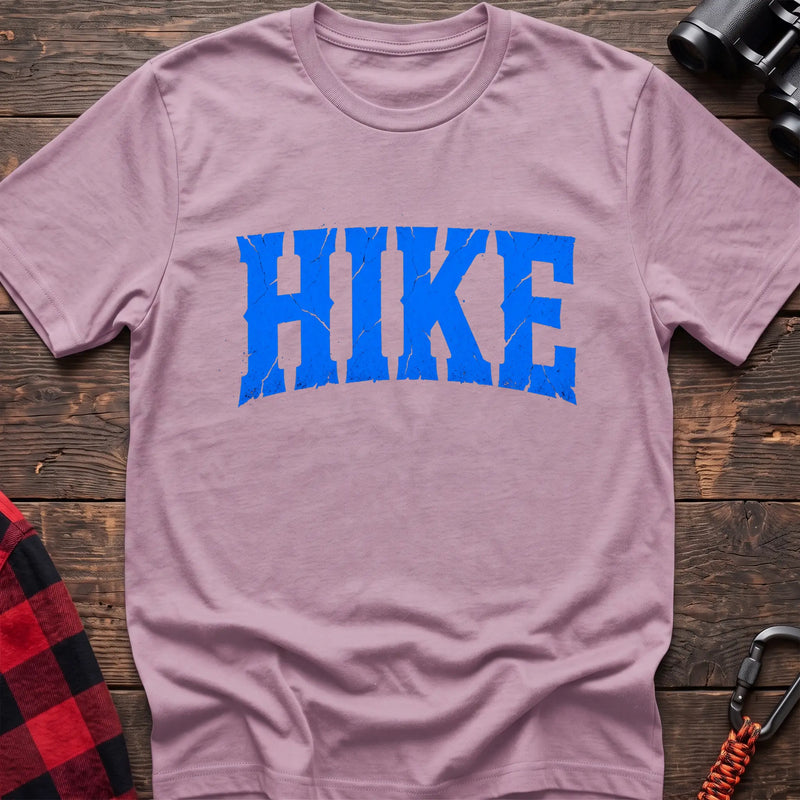 Light pink cotton t-shirt with distressed arched blue HIKE graphic in cracked varsity lettering, flat lay on rustic wood with hiking gear