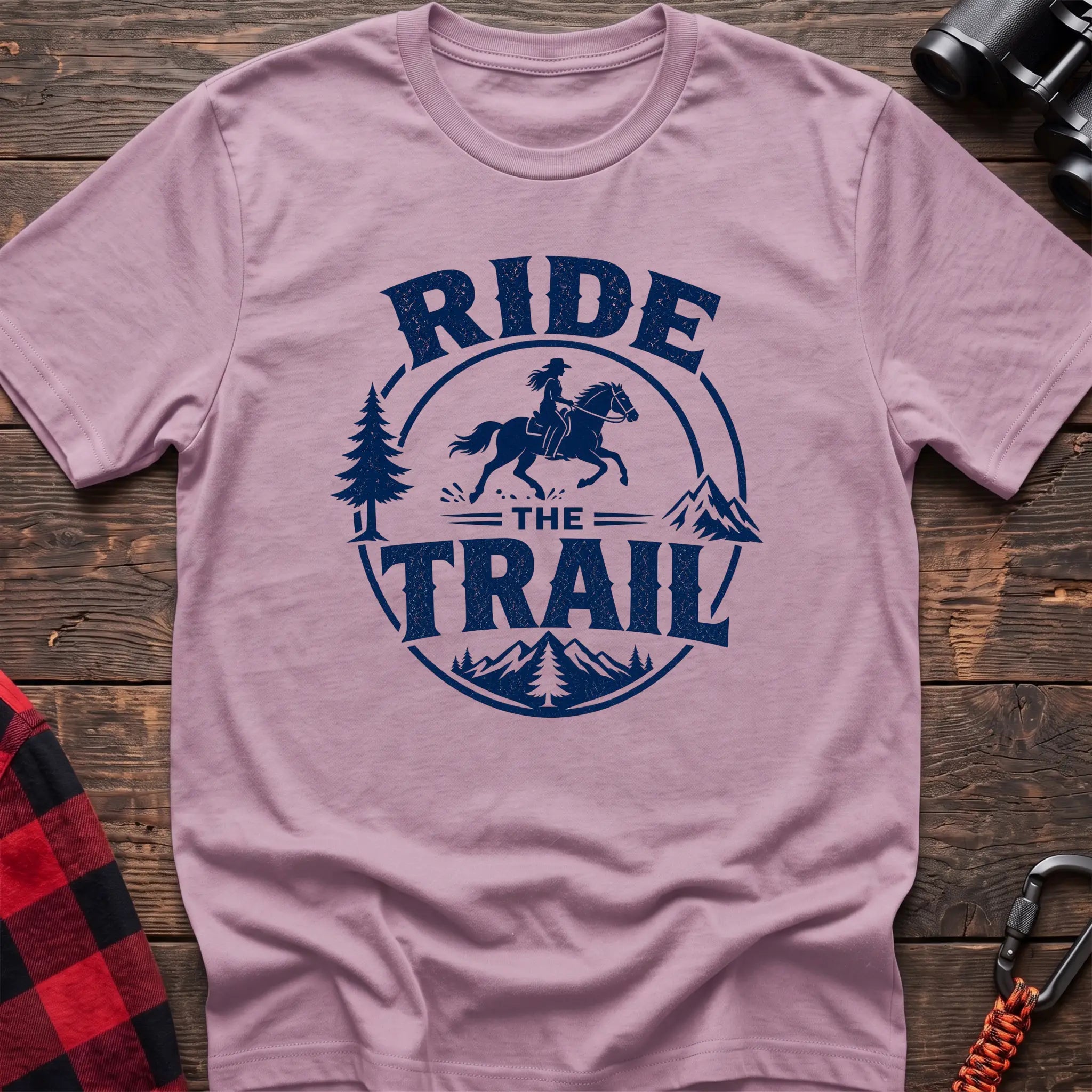 Light pink cotton t-shirt with navy Ride the Trail graphic of a woman horseback riding through mountains and pine trees, flat lay on rustic wood with hiking gear