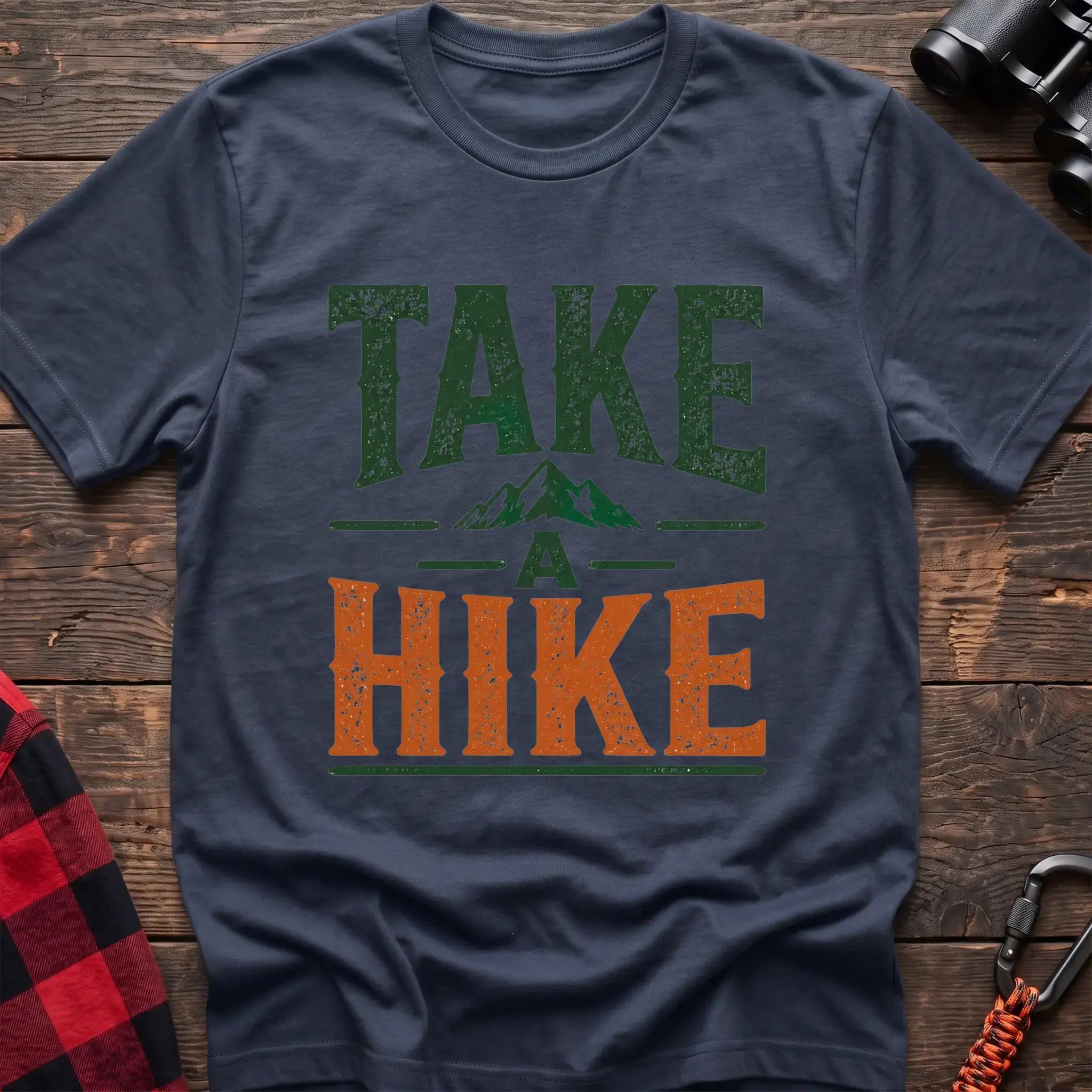 Navy t-shirt with distressed green and orange text reading “TAKE A HIKE” with a small mountain graphic between the words, laid flat on a wooden surface.