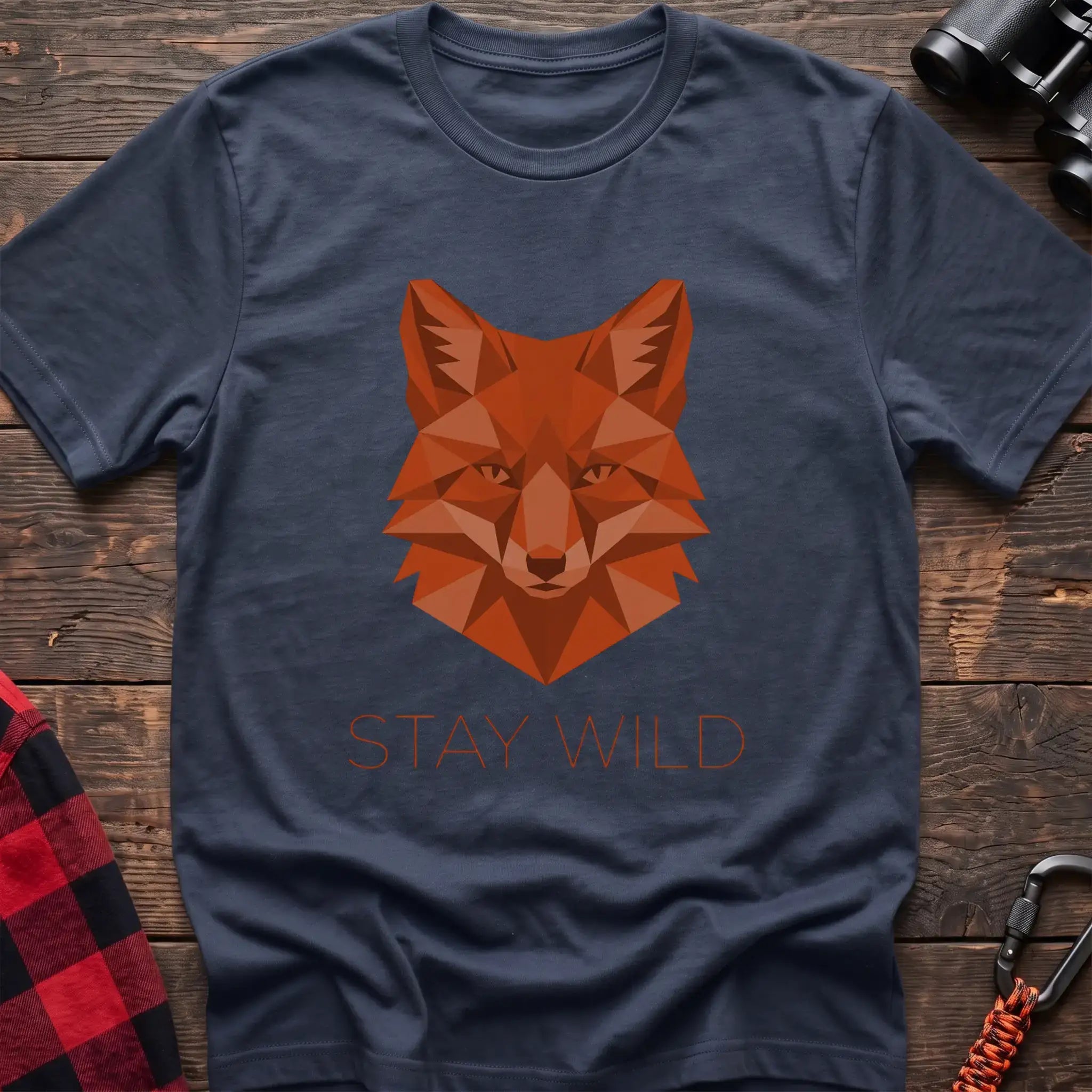 Navy t-shirt with a geometric low-poly orange fox head and the text “STAY WILD” printed below in matching orange, laid flat on a wooden surface.