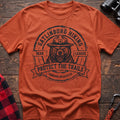 Orange cotton t-shirt with vintage Gatlinburg Hiking Bear League graphic of a roaring bear in a park ranger hat, Protect the Trails Tennessee Wildlife Division badge, flat lay on rustic wood with hiking gear