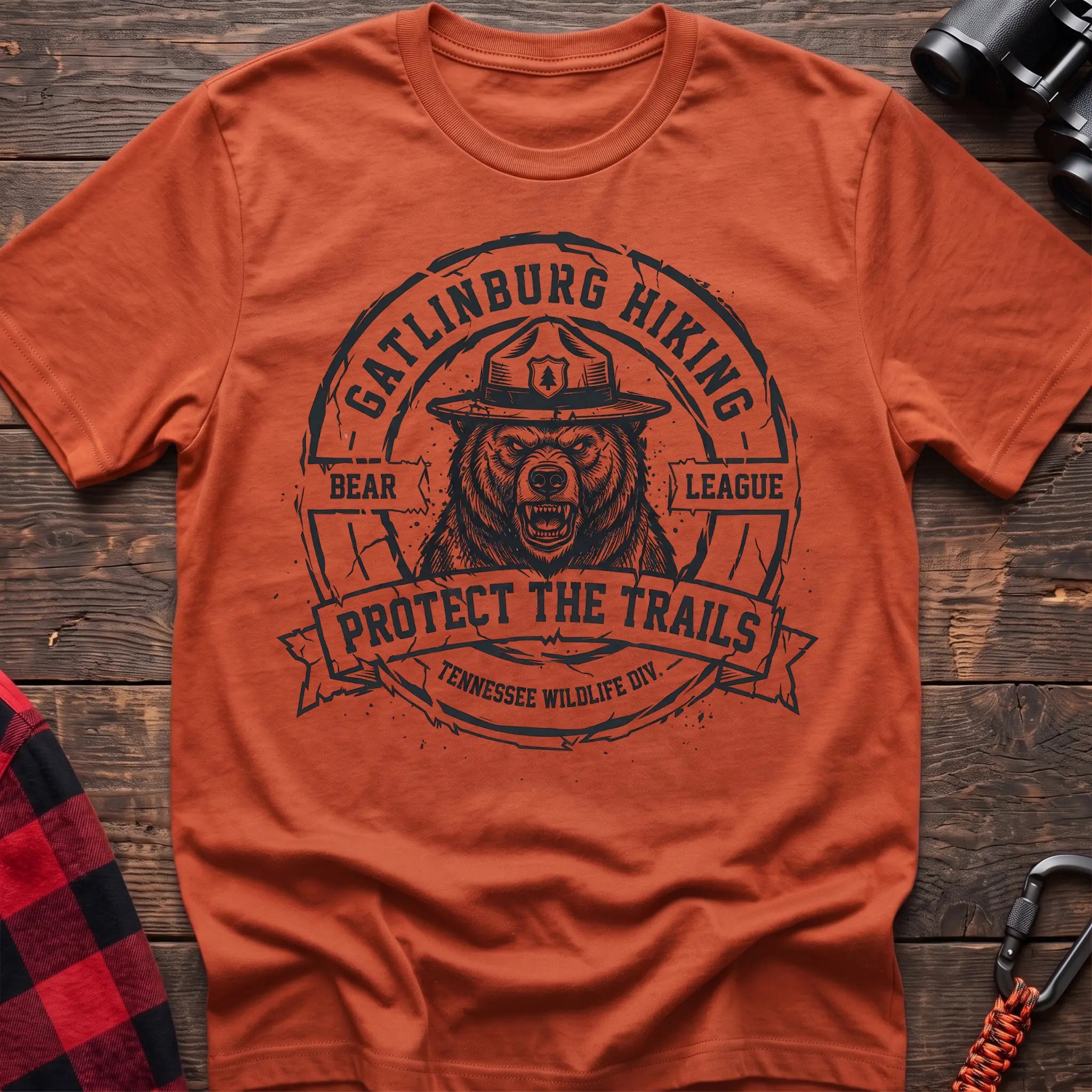 Orange cotton t-shirt with vintage Gatlinburg Hiking Bear League graphic of a roaring bear in a park ranger hat, Protect the Trails Tennessee Wildlife Division badge, flat lay on rustic wood with hiking gear