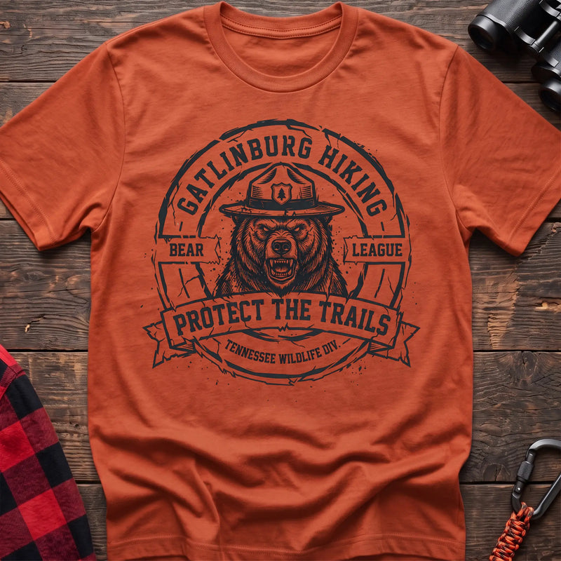 Orange cotton t-shirt with vintage Gatlinburg Hiking Bear League graphic of a roaring bear in a park ranger hat, Protect the Trails Tennessee Wildlife Division badge, flat lay on rustic wood with hiking gear