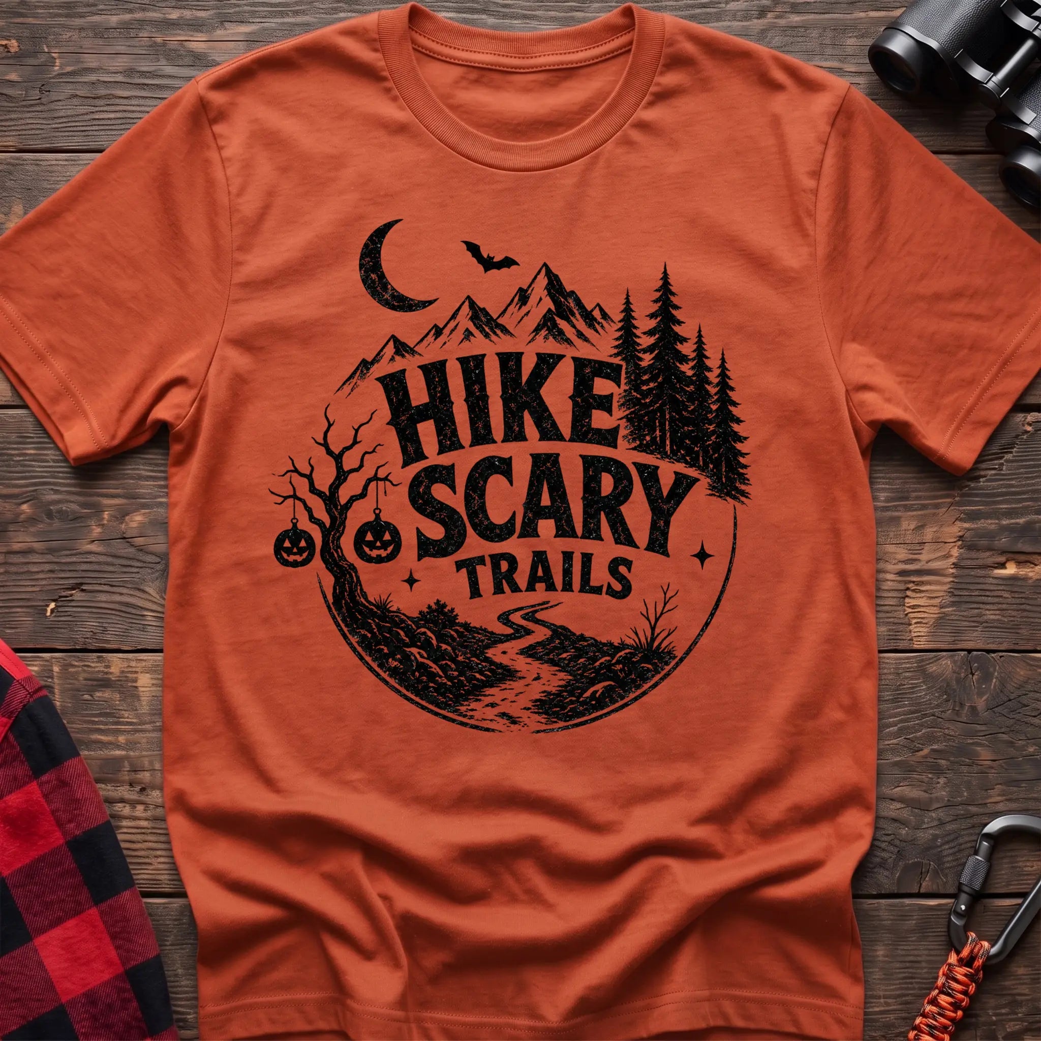 Orange cotton t-shirt with vintage Hike Scary Trails graphic of mountains, a crescent moon, bat, jack-o-lanterns, and a winding trail, flat lay on rustic wood with hiking gear