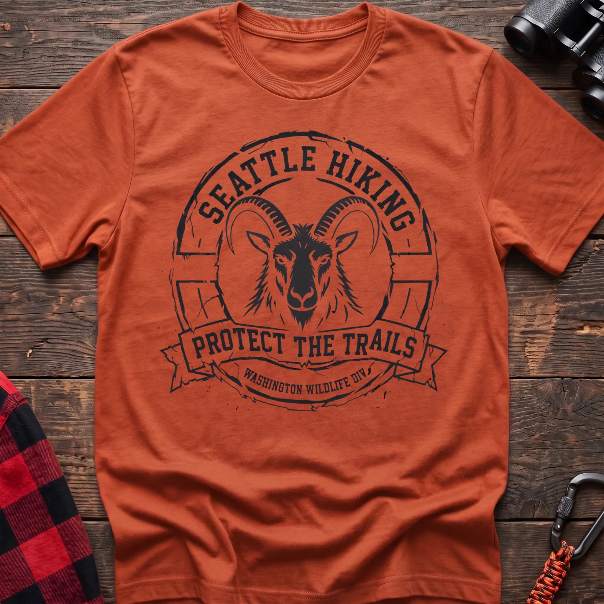 Orange cotton t-shirt with vintage Seattle Hiking graphic of a mountain goat in a circular badge, Protect the Trails Washington Wildlife Division design, flat lay on rustic wood with hiking gear