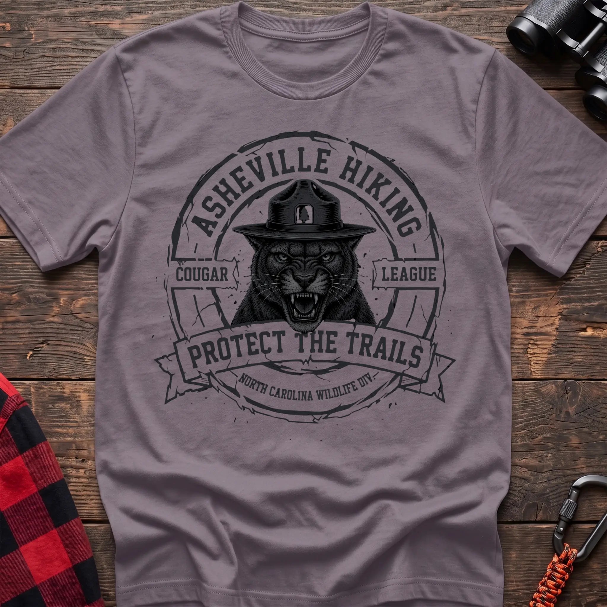 Paragon cotton t-shirt with vintage Asheville Hiking Cougar League graphic of a roaring cougar in a park ranger hat, Protect the Trails North Carolina Wildlife Division badge, flat lay on rustic wood with hiking gear