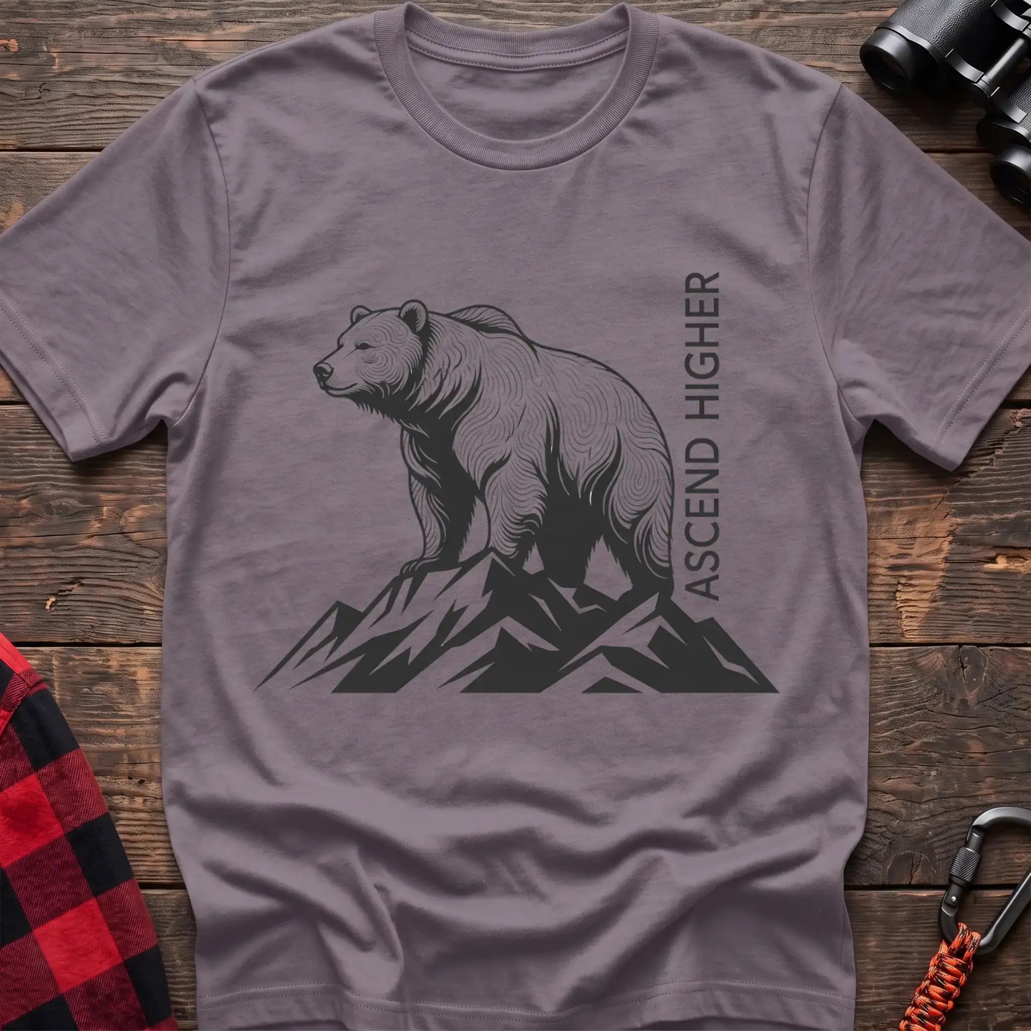 Paragon t-shirt with a black illustration of a bear standing on mountain peaks, with the vertical text “ASCEND HIGHER” beside it, laid flat on a wooden surface.