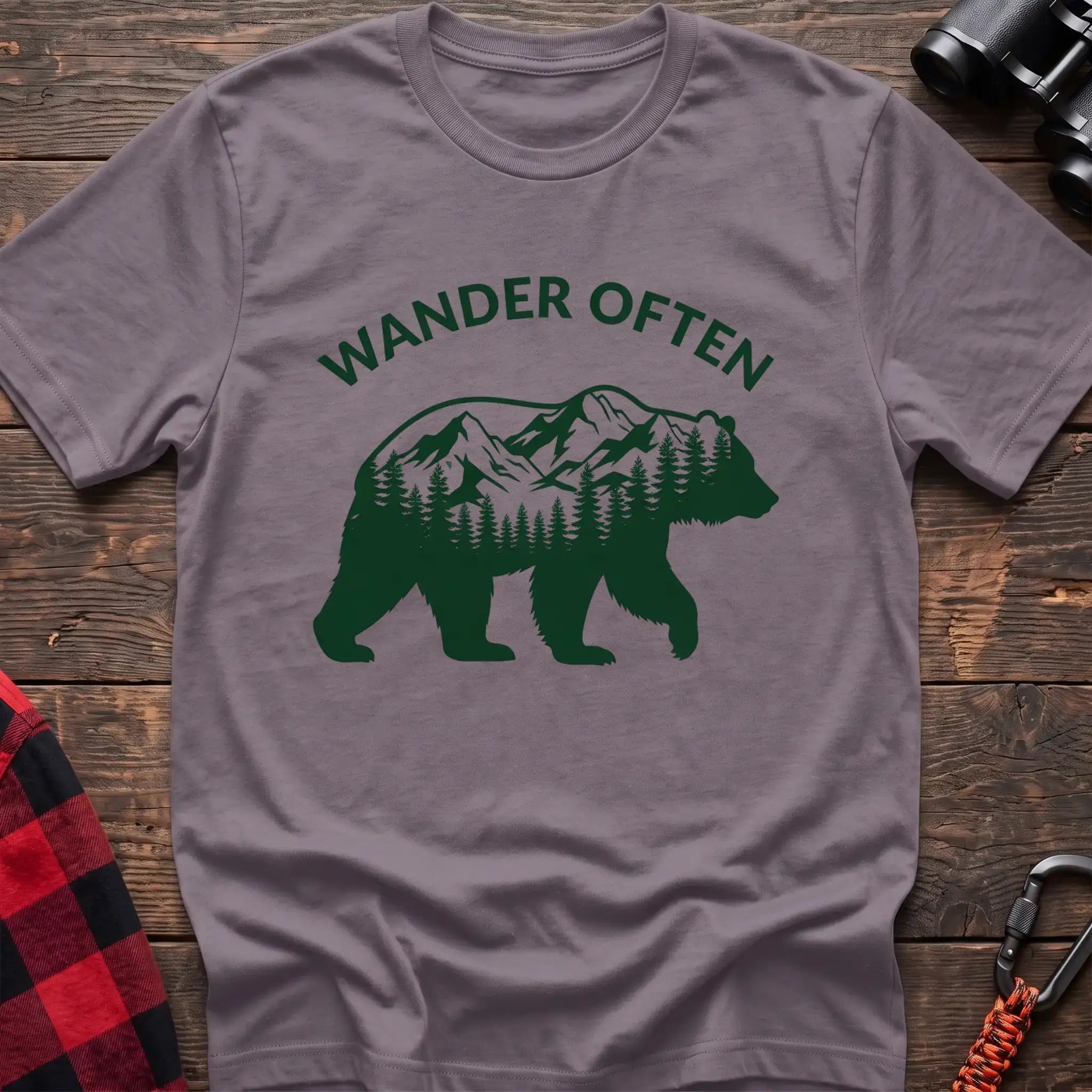 Paragon t-shirt with a dark green silhouette of a walking bear containing a mountain and pine forest scene inside, with the arched text “WANDER OFTEN” above it, laid flat on a wooden surface.